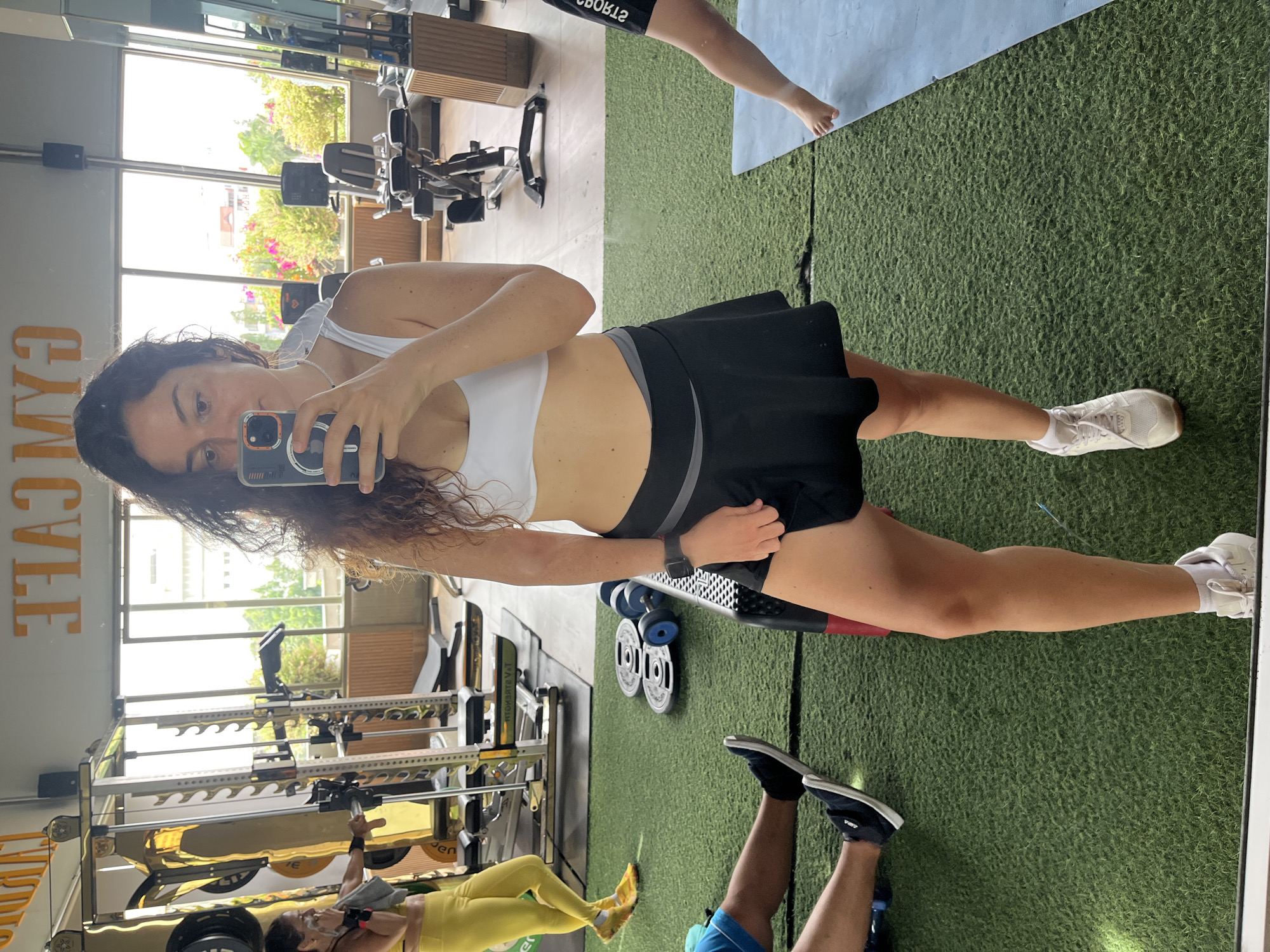 A woman stands in a gym, taking a mirror selfie. She is wearing a white sports bra and a black skirt with a grey waistband. Her left hand holds her phone, and her right hand is resting on her skirt. Her hair is curly and brown, falling over her shoulders. In the background, gym equipment is visible, including weight racks and exercise machines. Other people are also present in the gym, including a woman in yellow leggings to the left and two individuals with their legs on the floor to the bottom left. The gym has large windows that allow natural light to enter, revealing an outdoor scene with foliage. The floor is covered in green artificial turf. The signage above the windows reads "GYM CAFE" in large orange letters. The location is identified as the city of Phan Thiết, Vietnam.