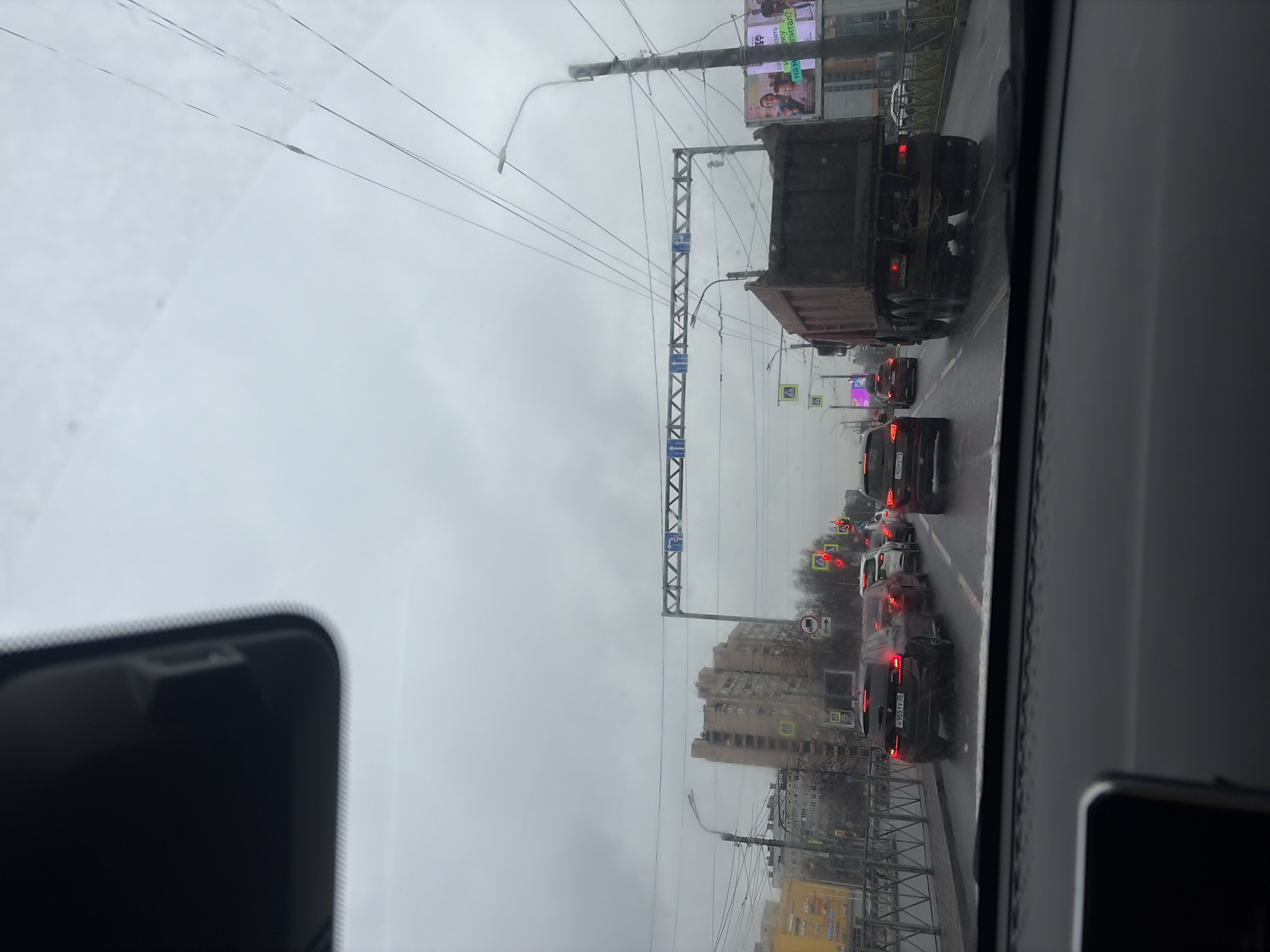 The image shows a view from inside a car, likely on a street in Saint Petersburg, Russia, given the Cyrillic license plate on a vehicle in traffic. It is an overcast day. A line of vehicles is ahead, including a large dump truck, moving slowly in what appears to be moderate to heavy traffic. Buildings line the street. Overhead wires and gantries with road signs are visible. There are no people visible outside the cars.