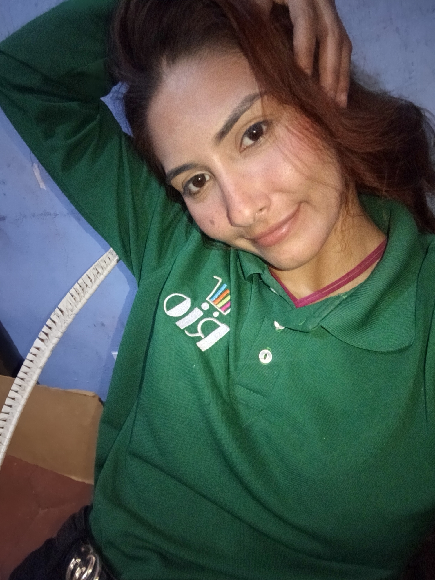 In Juan Griego, Venezuela, a young woman with auburn hair is pictured from the chest up, resting her head on her left arm. She is wearing a green polo shirt with a white logo on the left side, which appears to be the word "Bio" in stylized lettering with a colorful barcode-like design. A thin, dark pink ribbon is visible around her neck. Her right arm is bent at the elbow, with her hand resting on the back of her head, partially obscuring her hair. She is looking towards the camera with a slight smile. The background consists of a textured blue wall and a light-colored object with black markings, possibly a measuring tape or ruler, positioned to her left. The lighting suggests an indoor environment, possibly dim.