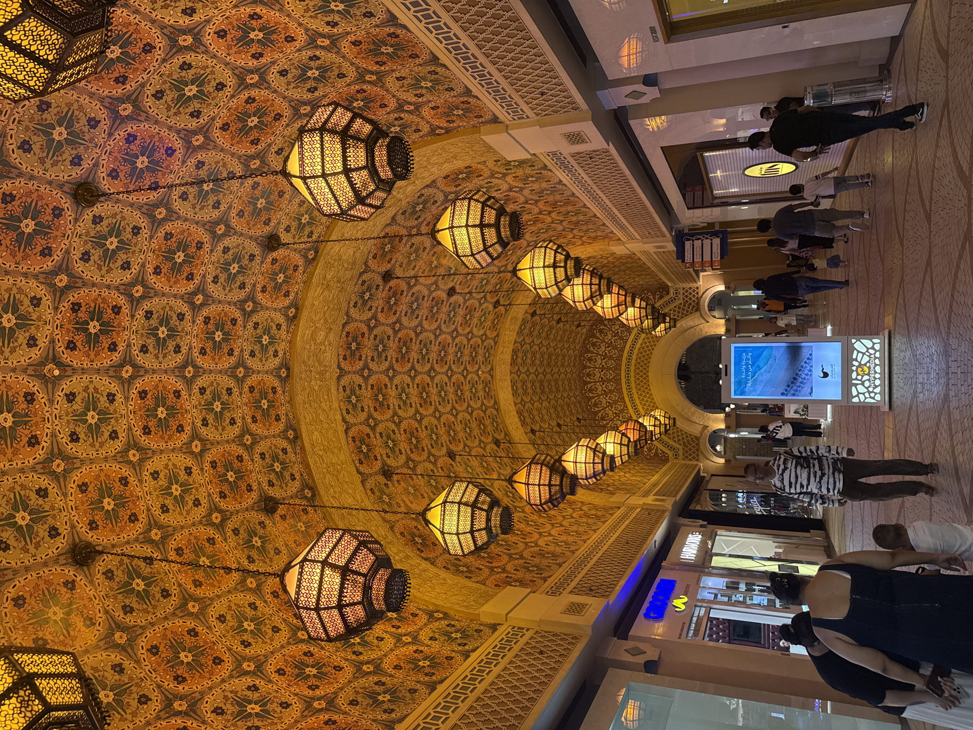 The image shows a bustling indoor shopping arcade, likely within the Souk Madinat Jumeirah in Dubai, United Arab Emirates, given its distinctive architecture and decorative elements. The ceiling is a vibrant tapestry of intricate geometric patterns in shades of orange, yellow, and blue, reflecting a traditional Islamic design aesthetic. Suspended from the ceiling by chains are numerous ornate Moroccan-style lanterns, casting a warm, inviting glow throughout the passageway.
The arcade features shops with illuminated storefronts on either side, their displays partially visible. In the foreground, several individuals are walking and interacting. On the left, a woman in a dark blue dress and a woman in a black top are facing each other, seemingly engaged in conversation. A young child in a white shirt stands between them. To their right, a man in a black and white striped long-sleeved shirt and dark pants walks away from the camera. Further down the arcade, more people are seen walking in various directions, indicating a lively atmosphere. A digital display board is prominent in the center, showcasing information or advertising. The floor is paved with textured tiles, creating a mosaic-like appearance. The overall impression is one of an aesthetically rich and commercially active public space, characteristic of Dubai's unique blend of modern amenities and traditional influences.