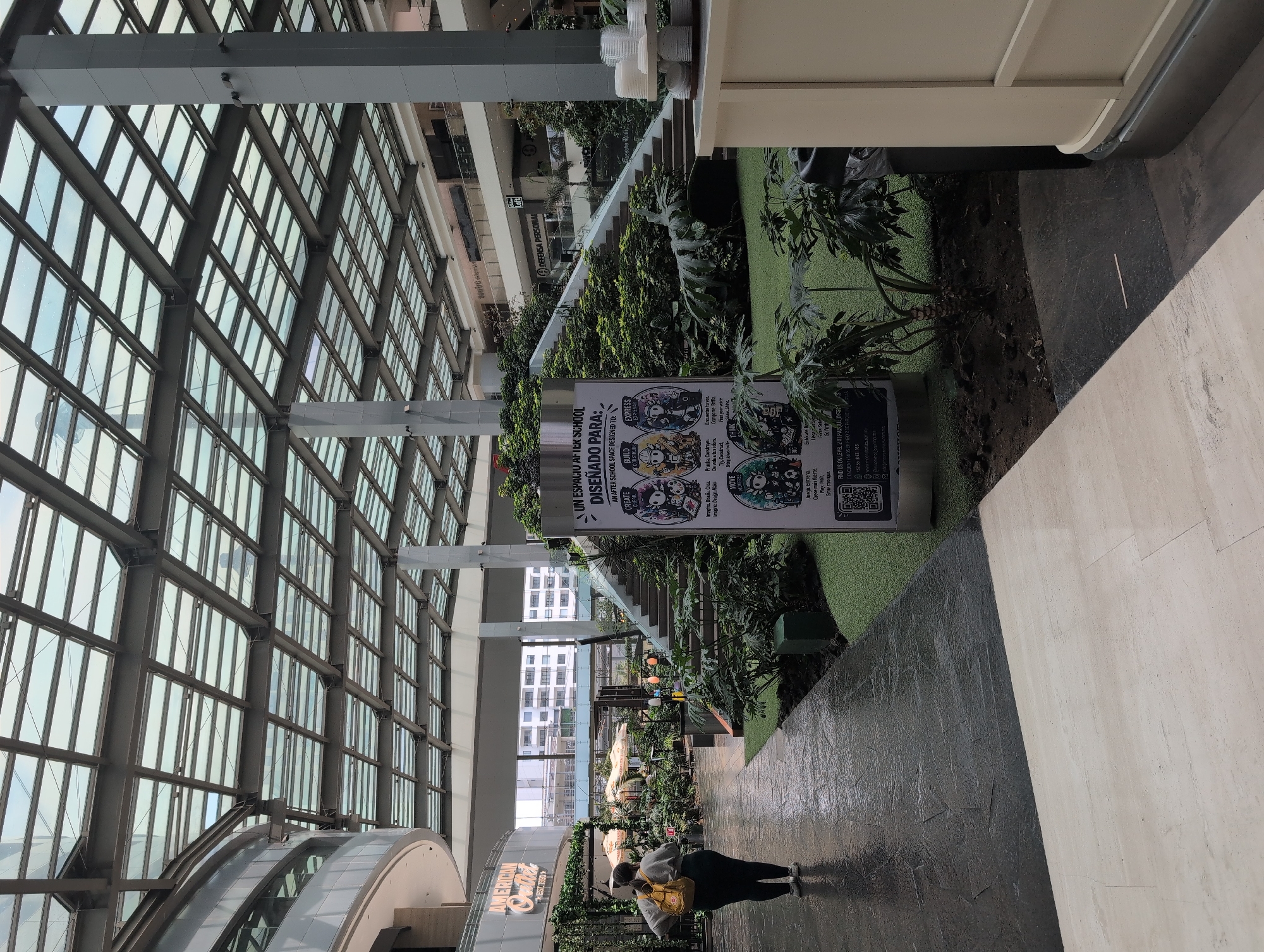 The image depicts an interior view, likely of a shopping mall or commercial space, in Jesús del Monte, Mexico. The architecture features a large glass ceiling supported by metal framework, which allows for ample natural light. An elevated walkway or second level is adorned with greenery, including various plants and artificial turf. A sign in Spanish, advertising an after-school program with the words "UN ESPACIO AFTER SCHOOL DISEÑADO PARA" and further text detailing "CREATE", "BUILD" and "EXPRESS", is prominently displayed. The floor is divided into dark and light sections. A person is walking on the darker section of the floor. Store signs are visible in the background, including "AMERICAN OUTLET" and "DEFENSA PERSONAL."