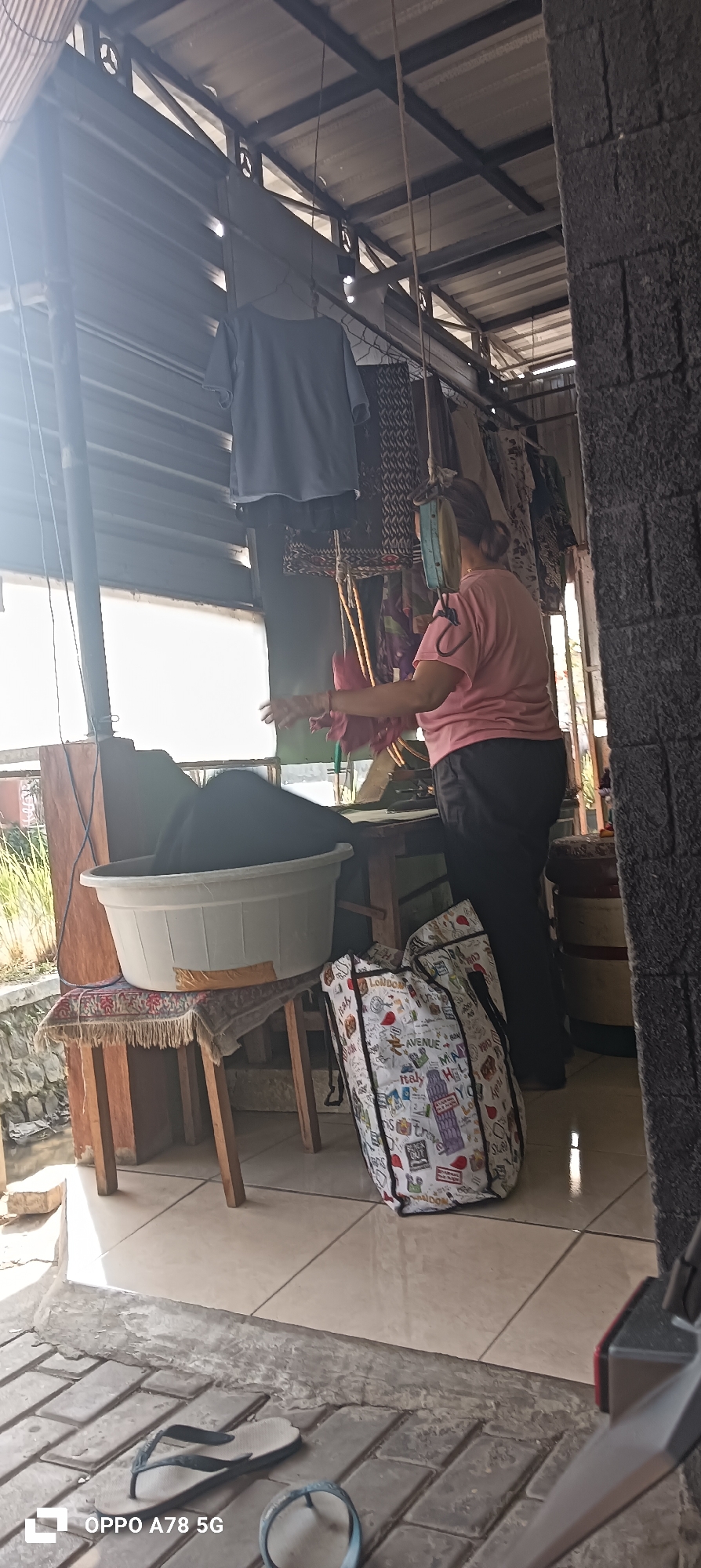 A medium shot captures an outdoor area in Buncitan, Indonesia, likely a laundry or preparation area, with a woman in a pink t-shirt and black pants in the center. She is facing away from the camera, reaching towards a large white plastic basin filled with dark fabric. Behind her, clothes hang from a line strung across a metal-framed structure with a corrugated metal roof. A patterned tote bag with images of landmarks like the Eiffel Tower and Big Ben rests on the tiled floor to her right. To her left, the basin is placed on a wooden stool covered with a patterned cloth. The environment appears to be an open-sided structure, possibly a patio or veranda, with a stone wall on the far right and a bright window or opening behind the basin. The floor is tiled with a concrete border. In the foreground, a pair of flip-flops lies on the tiled floor. The lighting suggests it is daytime, with sunlight casting shadows.