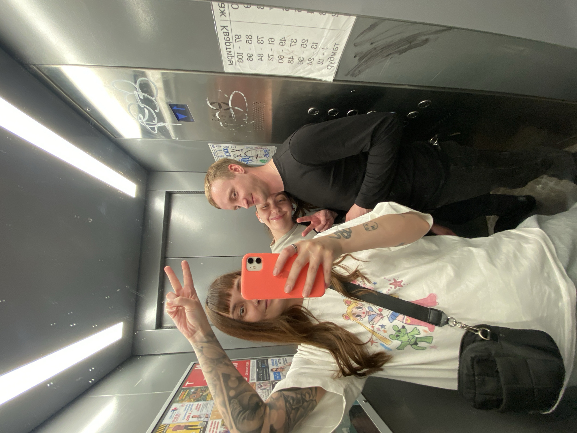A slightly rotated, medium shot shows three individuals in what appears to be an elevator in Votkinsk, Russia, based on the Cyrillic text on the wall. The focus is on the young woman in the foreground who is holding an orange cell phone to take a selfie in a reflective, stainless steel elevator. She is wearing a white t-shirt with cartoon characters printed on it, has reddish brown hair with bangs, and multiple tattoos are visible on her left arm. Her right hand is raised in a peace sign. A small black purse hangs on her right side.
Behind her, a man in a black long-sleeved shirt is seen. Next to him, a second woman appears, partially obscured by the first woman and the man.
Above the group, a sign written in Cyrillic is affixed to the elevator wall. The elevator interior features a strip of white light and a stainless steel finish. There are some graffiti marks on the elevator.