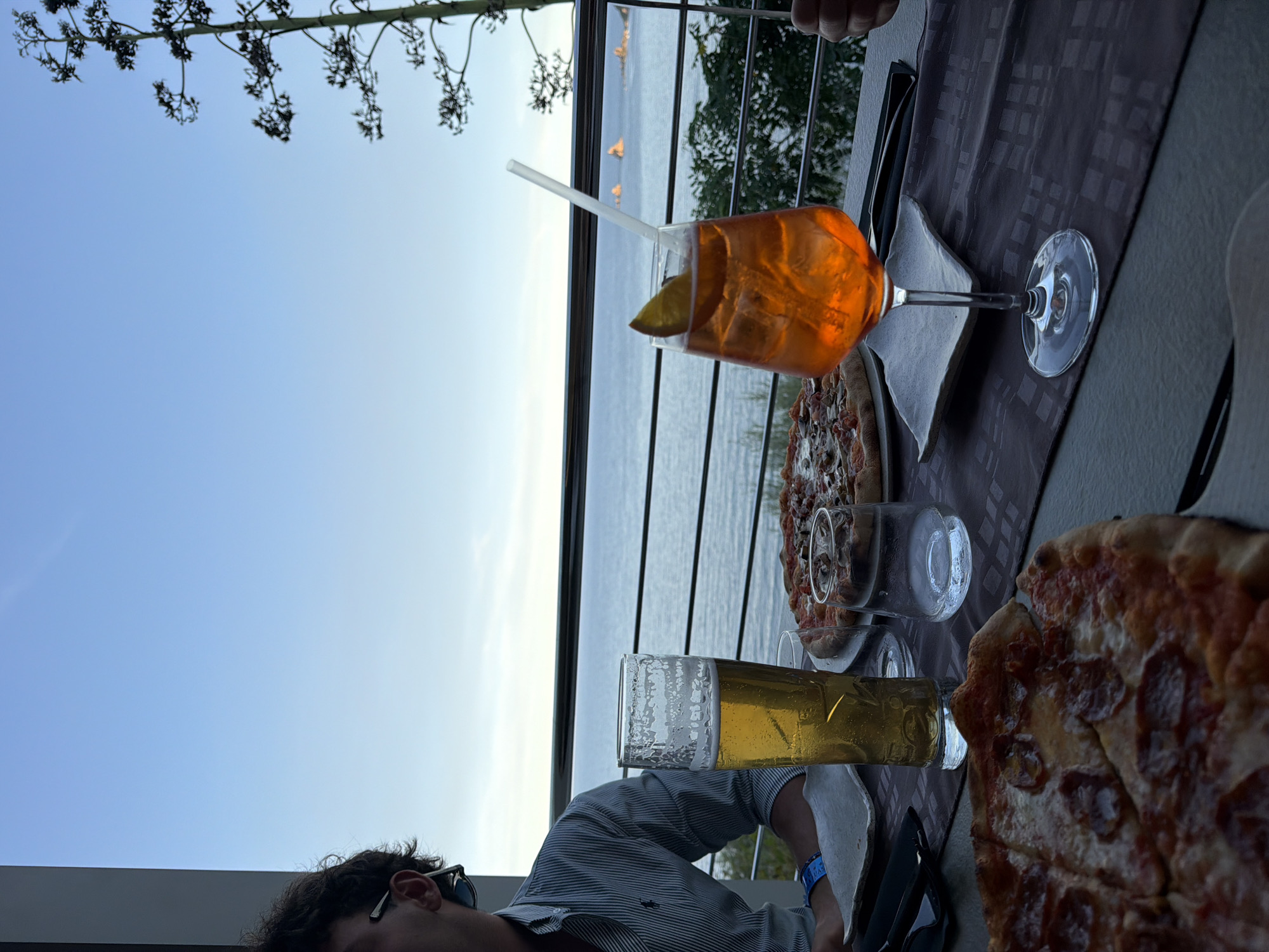 A close-up, eye-level shot shows a table set for a meal with a picturesque view of the sea and sky. In the foreground, a pepperoni pizza is partially visible, while a second pizza with mushrooms and cheese sits further back on the table. Two drinks are present: a tall glass of beer with foam and a vibrant orange cocktail garnished with a slice of orange and a white straw. A person, partially visible on the left, wears a striped shirt and sunglasses on their head. The table is covered with a dark-patterned tablecloth. In the background, a railing separates the table from the sea, which appears calm under a clear, pale blue sky. A tall, dark, spiky plant is visible on the right side of the frame, with the scene suggesting a coastal restaurant setting in Santa Maria Navarrese, Italy.