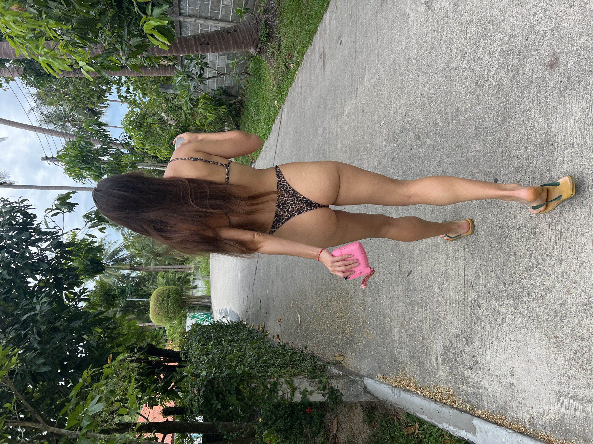 A full-length, rear-view shot shows a woman walking along a concrete path in a lush, tropical setting. She has long, dark brown hair extending down her back and is wearing a leopard print bikini, with the top straps visible over her shoulders and matching bottoms. In her left hand, she holds a small, bright pink water bottle or flask with a strap. She is wearing yellow or tan flip-flops with green straps on her feet.
The environment is verdant and sunny, characteristic of a tropical climate. On the left side of the concrete path, tall palm trees and various broadleaf trees and bushes create a dense green backdrop. On the right, the path is bordered by a low concrete curb, beyond which more green foliage and a grey brick wall are visible in the background. The concrete path appears well-maintained but has scattered light-colored debris or small leaves on its surface.
The sky, visible in glimpses through the trees, is light blue with some white clouds, indicating daytime, likely under bright conditions. The overall scene suggests a casual stroll in a warm, outdoor environment, aligning with the specified location of บ้านบางรักษ์, Thailand. There is no visible text in the image.