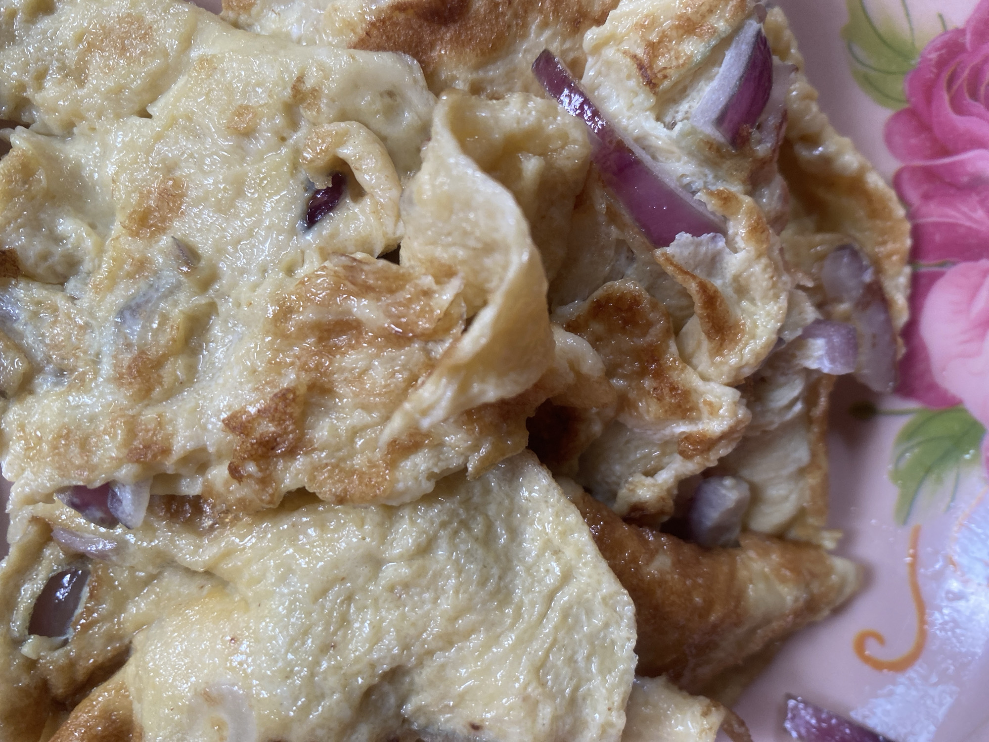 A close-up, overhead shot captures a pile of cooked eggs, possibly an omelet or scrambled eggs, mixed with pieces of red onion. The eggs are a pale yellow with some areas browned from cooking. The red onion pieces are visible as purple slivers interspersed throughout the eggs. The food is presented on a pink plate adorned with floral designs, featuring pink roses and green leaves. The lighting is bright, casting a slight sheen on the eggs. The image does not contain any information about the city of Lafia, Nigeria or any people or actions.