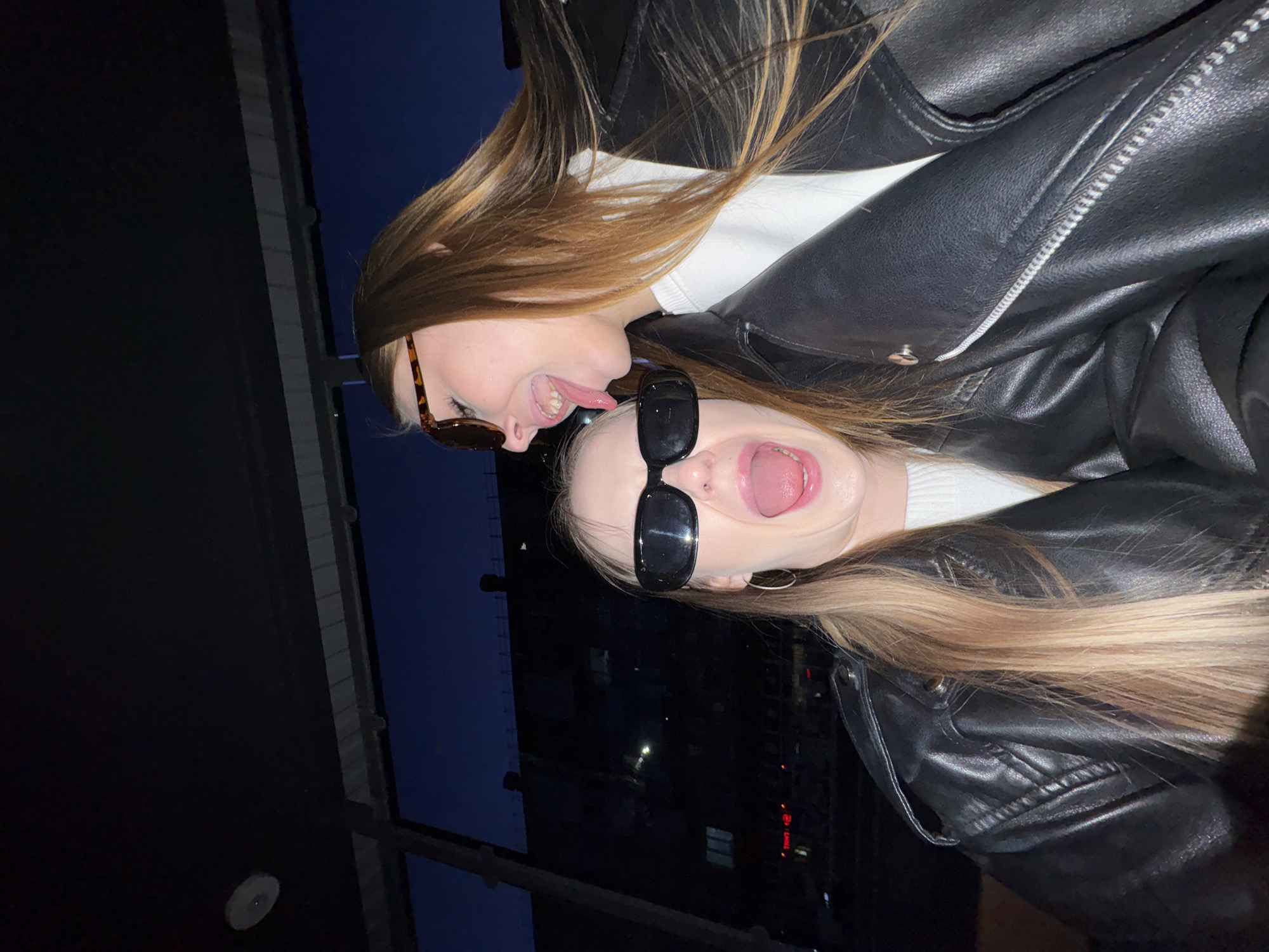 Two young, light-skinned women wearing black leather jackets and sunglasses are posing in what appears to be an outdoor setting, likely a balcony or rooftop. The woman on top is wearing tortoise-shell sunglasses and has her tongue extended towards the bottom woman. The woman below has black sunglasses and is shouting with her mouth open. The city of Chaikovsky, Russia is visible in the background.