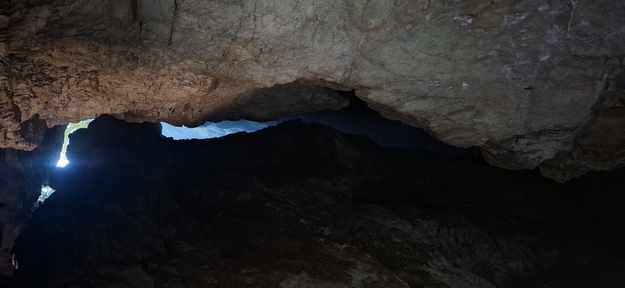 The image shows a view looking upwards into a narrow, dark crevice within a rocky formation, likely a cave or canyon. Sunlight streams in from an opening at the top, illuminating the jagged edges of the rock. The rock faces are textured and appear to be made of limestone or a similar sedimentary rock, with variations in color from light brown to grey. The crevice itself plunges downwards into darkness, with glimpses of a bright blue sky or distant opening visible in the depths. The scene suggests a dramatic geological feature, possibly a natural bridge or a narrow passage through a cliff.
This specific view is characteristic of the Trigrad Gorge in Bulgaria, known for its dramatic karst landscape and natural formations like the Devil's Throat Cave and the Trigrad Gorge itself. The deep fissures and narrow passages carved by water over millennia are a hallmark of this region.