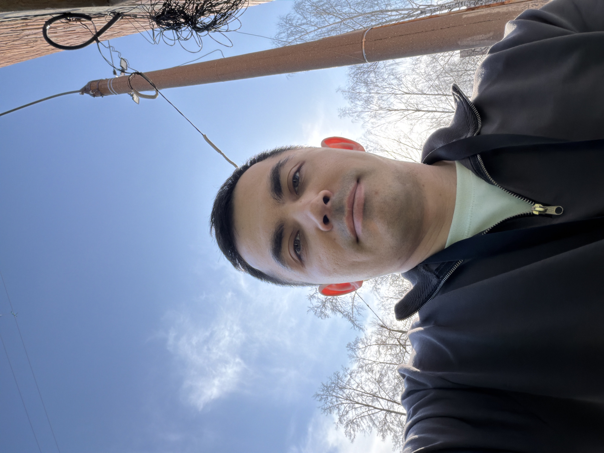 A low-angle, selfie-style photograph shows a man wearing a black jacket and a light-colored t-shirt against a background of a partly cloudy blue sky. Bare tree branches are visible in the upper right and lower center of the frame, indicating winter or early spring. Above the man and slightly left of center, a utility pole and various wires are visible. Based on the presence of utility poles and bare trees in an urban setting under a partly cloudy sky, the location might be Izhevsk, Russia. The photo focuses on the man's face, with the camera positioned slightly below eye level.