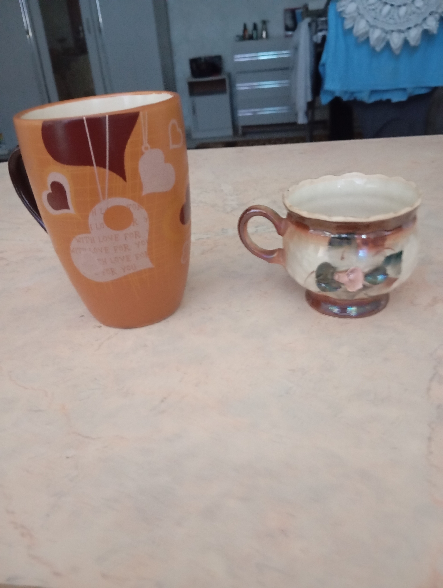 Two ceramic mugs sit on a light-colored, marbled surface. The mug on the left is a tall, orange mug with a dark brown handle. It is decorated with dark brown and white hearts, and the text "WITH LOVE FOR YOU" repeated in white. The mug on the right is smaller and wider, with a decorative brown glaze that fades to white. It features a painted pink flower with green leaves. The background is blurred, but appears to be an indoor setting with furniture and clothing visible, suggesting a domestic environment in Strășeni, Moldova.