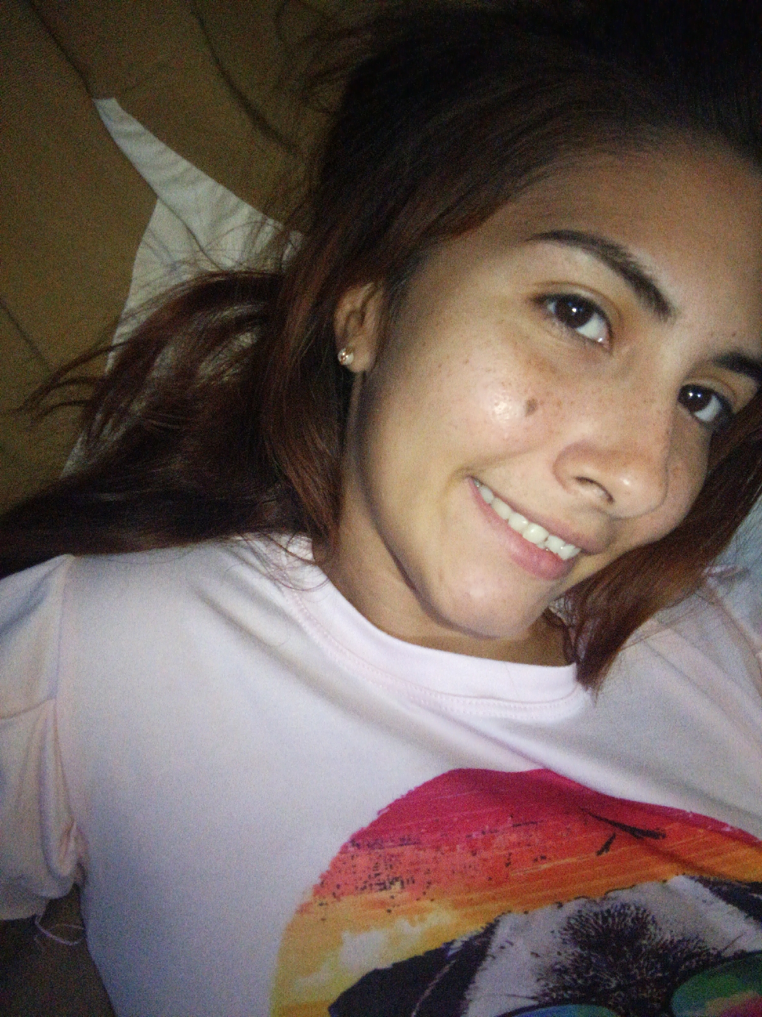 The provided image is a close-up, indoor selfie of a young woman. She has brown hair, fair skin with freckles, and is wearing a light pink t-shirt with a graphic print. She is smiling, showing her teeth, and looking directly at the camera. The background appears to be a bed or couch with a brown textured surface and a white sheet or pillow. The image quality is grainy, suggesting it was taken in low light conditions. There is no visible indication of the location being in Juan Griego, Venezuela, or any other specific geographical area. The scene is a personal, informal moment captured by the subject herself.