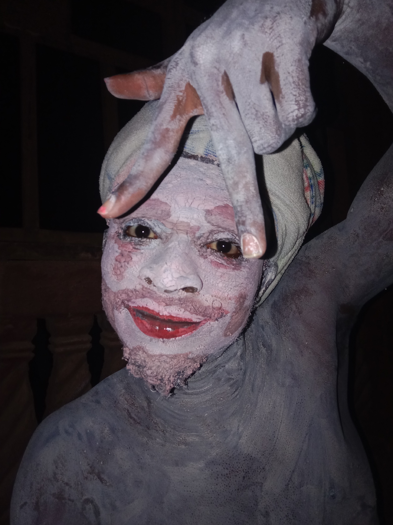 A person is covered in white paint on the face and body, with red lipstick applied to the lips. The person is wearing a cloth wrapped around their head and is raising one arm with the hand in a gesture above the head. The background is dark, with the vague outline of a wooden structure visible.