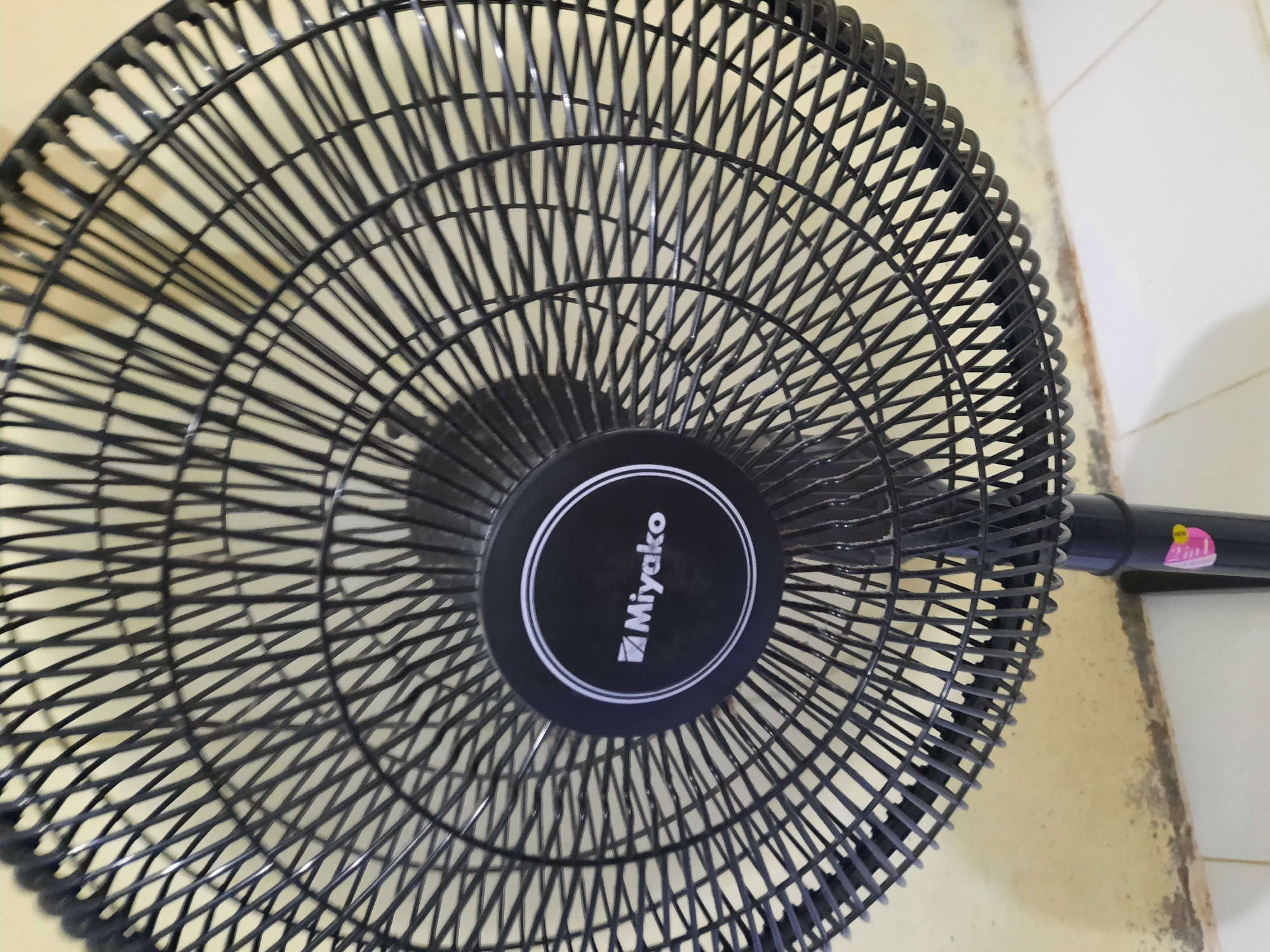 The image shows a close-up of a black electric fan with the brand name "Miyako" visible on a circular emblem in the center of the fan's grill. The fan is positioned in front of a light-colored, textured wall, possibly a domestic interior in the city of Lhokseumawe, Indonesia. The fan's grill is made of thin, parallel metal bars, forming a dense mesh. The stand of the fan is partially visible at the bottom, and there is a sticker with pink and yellow elements on it. The floor below appears to be tiled in white.