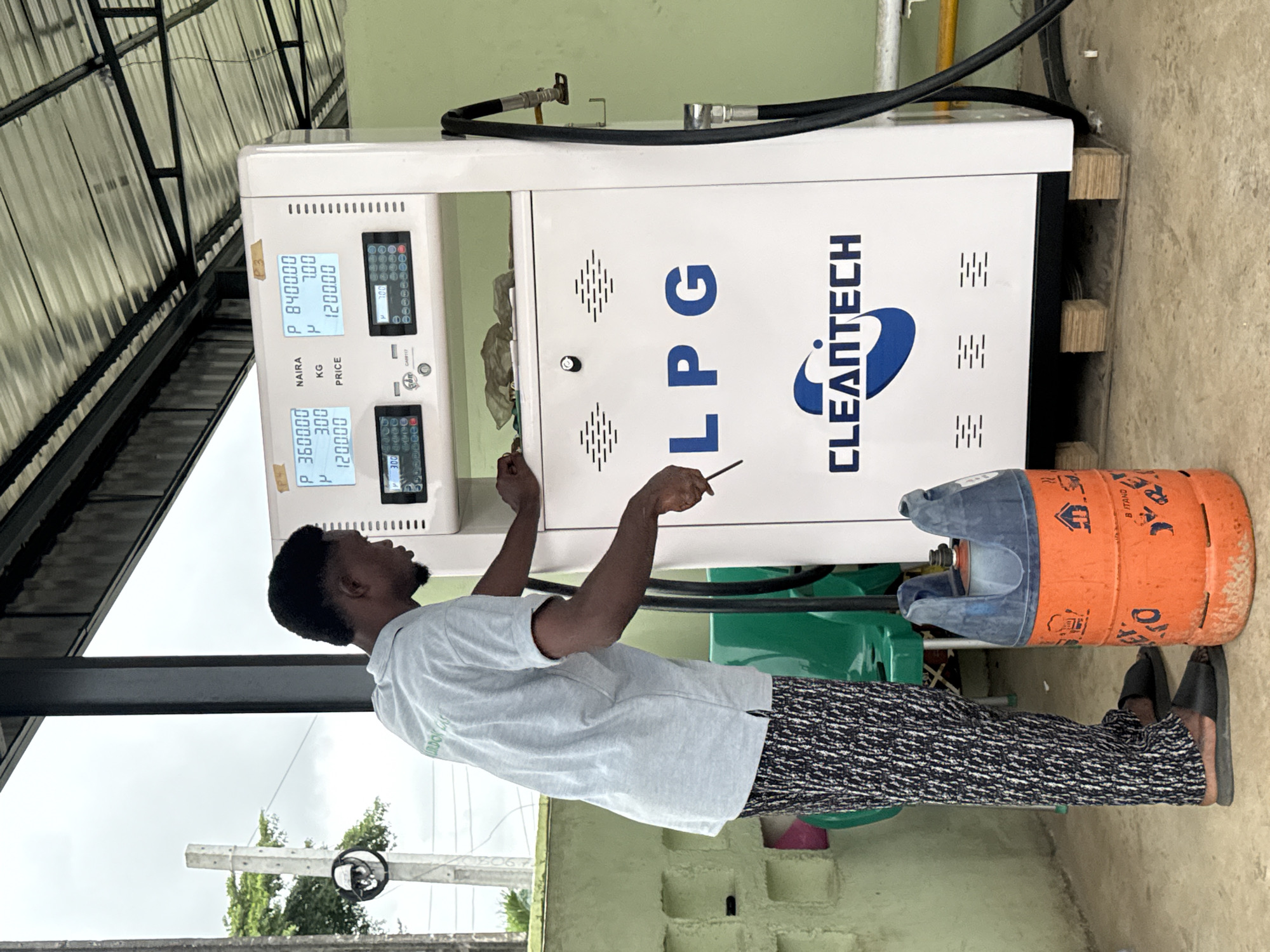 In Ado, Nigeria, a man is seen standing next to an LPG dispensing pump. He is wearing a light blue polo shirt and black and white patterned trousers. He appears to be connecting a hose from the pump to an orange gas cylinder. The LPG pump is white with a digital display showing price, kilograms, and quantity. The brand name "CLEANTECH" is visible on the pump. The scene is outdoors, under a covered structure with a metal roof. A concrete wall is visible in the background, along with some greenery.