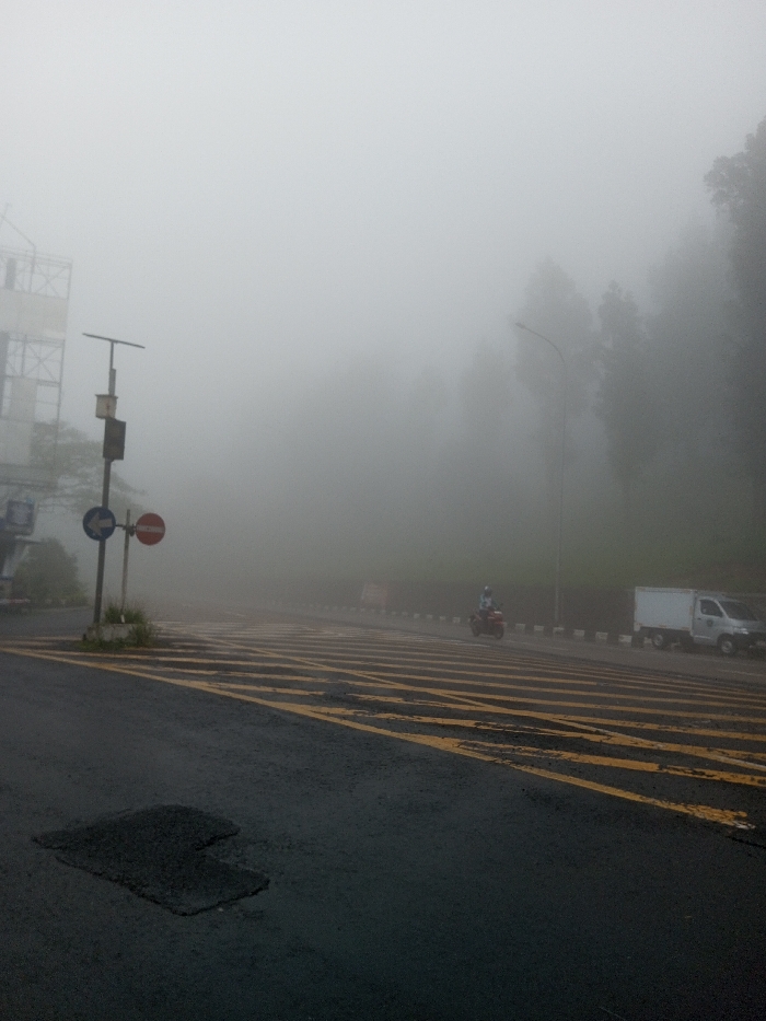 A foggy road scene with yellow stripes across the asphalt. A street sign with a no-entry symbol stands to the left. In the distance, a motorcyclist rides alongside a small white delivery truck. Trees and a building are visible through the thick fog. The overall atmosphere is muted and visibility is greatly reduced due to the heavy fog.