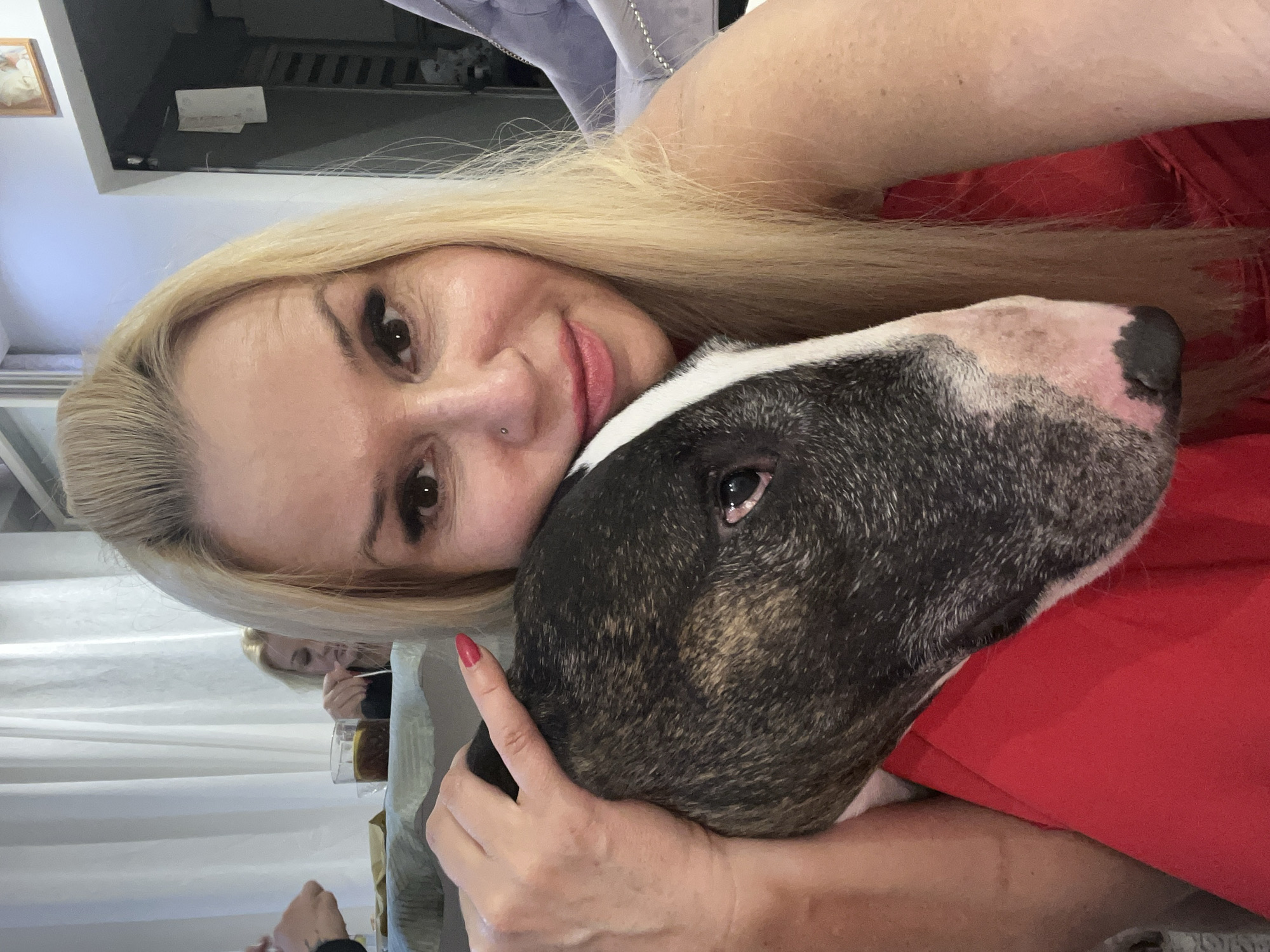 In a selfie taken in Białystok, Poland, a woman with blonde hair and a red dress poses with a bull terrier. The woman's left arm is around the dog, which rests its head on her shoulder. The background shows a curtain and furniture.