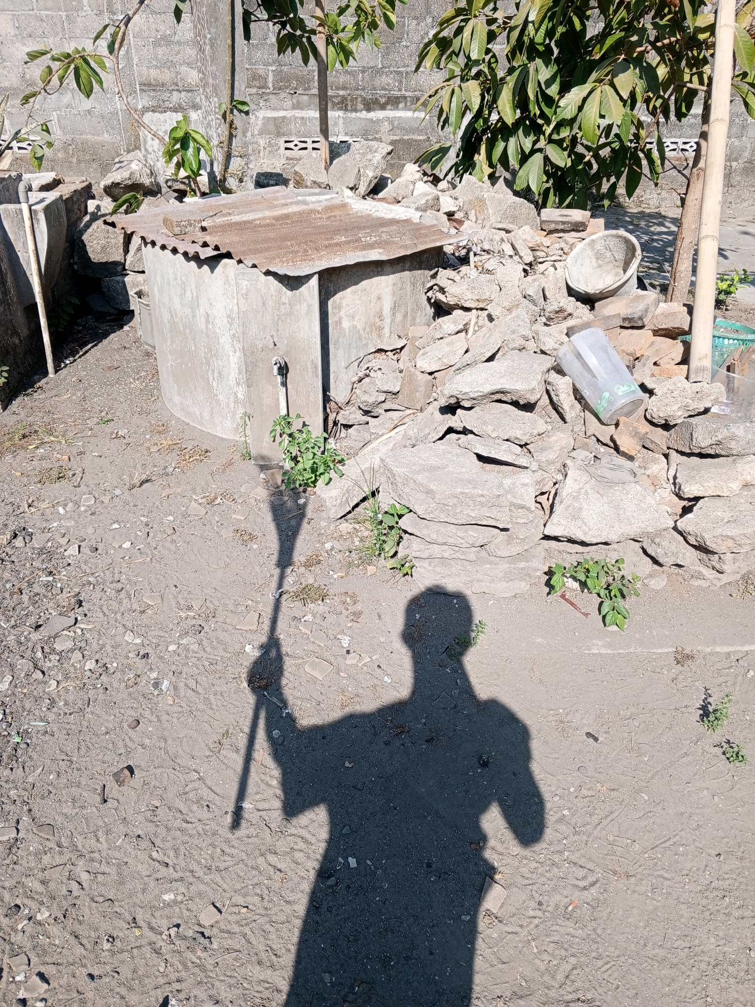 The image depicts an outdoor scene in the city of Bantul, Indonesia. In the foreground, a shadow of a person holding a long object, possibly a stick or a pole, is cast on sandy ground with scattered debris. The ground appears to be a mixture of dirt, small stones, and fragments of broken objects.
In the middle ground, there is a concrete structure that resembles a well or a water tank, covered with a rusty corrugated metal roof. A spigot or tap is visible on the side of the concrete structure. To the right of the concrete structure, a pile of large, irregularly shaped stones and bricks is present. Among the stones, there is a clear plastic container, possibly a pitcher, and a small, light-colored basin. A thin bamboo pole stands vertically near the pile of stones.
In the background, a rough brick wall forms a backdrop. A tree with green leaves is positioned above and behind the concrete structure and the pile of stones. The overall environment suggests an outdoor area, possibly a yard or a vacant lot, with elements of construction and debris. The lighting indicates a sunny day, with the sun casting a distinct shadow.