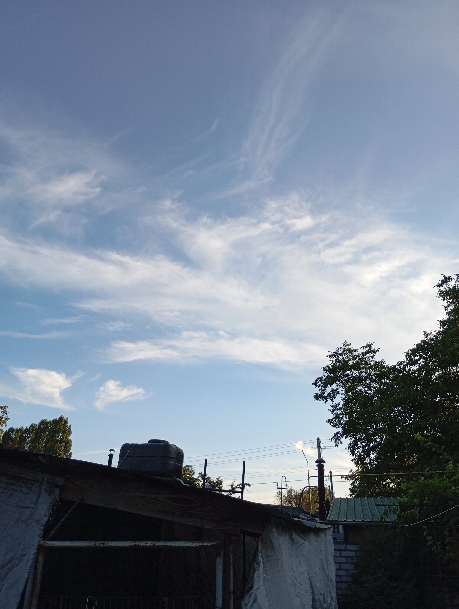 The image depicts an outdoor scene, likely in a residential or industrial area, with a focus on the sky and structures below. The sky is a pale blue with wispy white cirrus clouds stretching across the frame, suggesting daylight with some atmospheric activity.
In the foreground, partially obscuring the view, is a rustic structure with a sloped, dark-colored roof. A large, black plastic water tank is visible on top of this structure. The structure itself appears to be made of wood and covered in a light-colored tarp or sheeting, suggesting it might be a shed or an unfinished building.
Behind this main structure, a dense green tree with full foliage dominates the right side of the image. To the left of the tree and slightly behind the main structure, a line of tall, slender trees with a more sparse canopy is visible.
Further into the background, utility poles with multiple wires strung between them are present. There is also a glimpse of a green corrugated metal roof on another building to the right. The overall impression is one of a functional, perhaps somewhat unkempt, outdoor space.
The image does not contain any people or any discernible actions or interactions. The scene is static and environmental. The city of Horishni Plavni, Ukraine, is mentioned, implying a geographical context, but there are no specific landmarks within the image that would definitively confirm this location without external information. The lighting suggests it is likely morning or late afternoon, given the direction of the light and the quality of the sky.