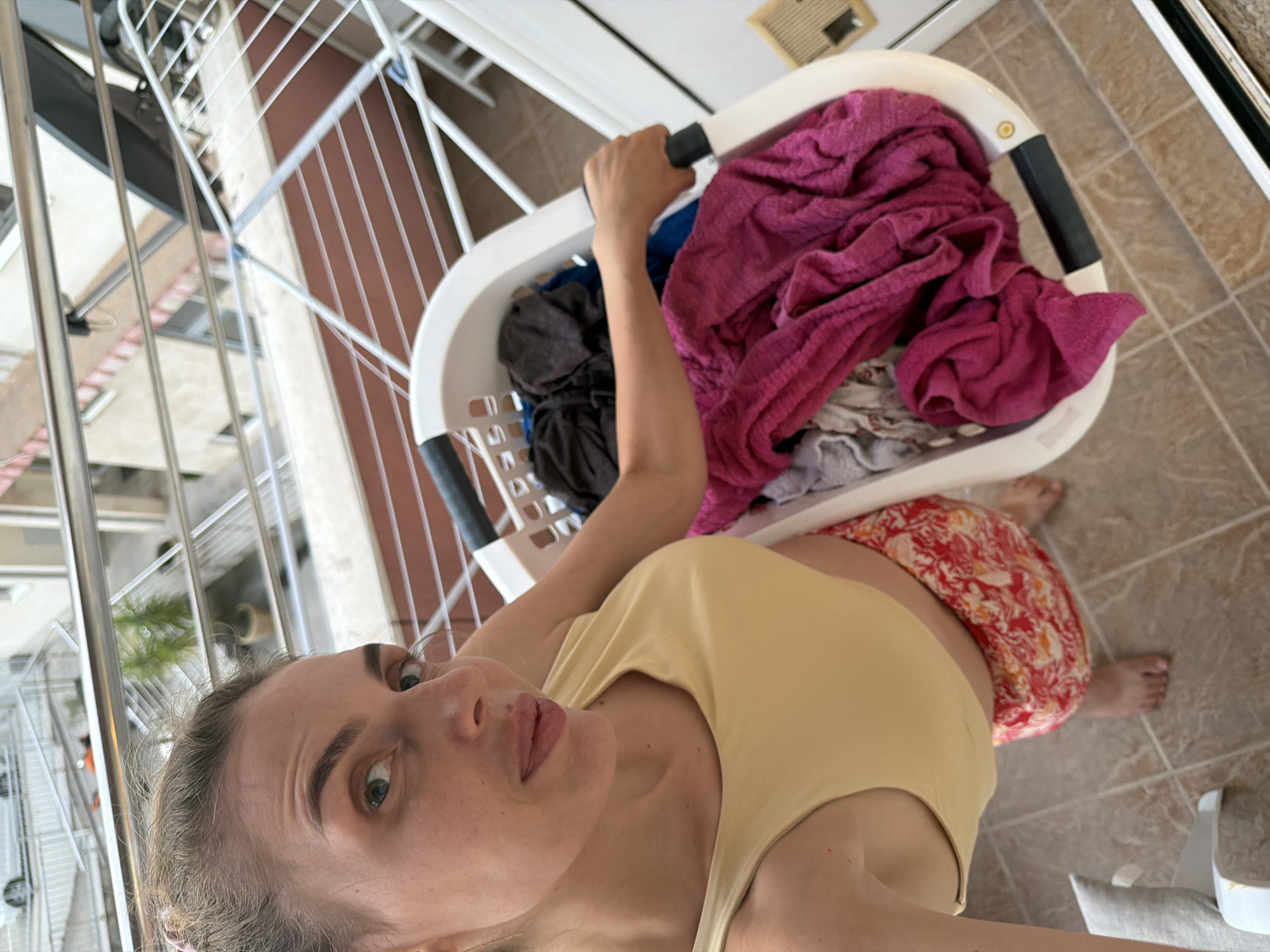 In Budva, Montenegro, a woman stands on a tiled balcony, holding a laundry basket filled with clothes. She is wearing a yellow crop top and a colorful skirt. Behind her, a drying rack is visible, and beyond that, the exterior of a building and a street with a car. The scene appears to be outdoors, possibly on a sunny day.