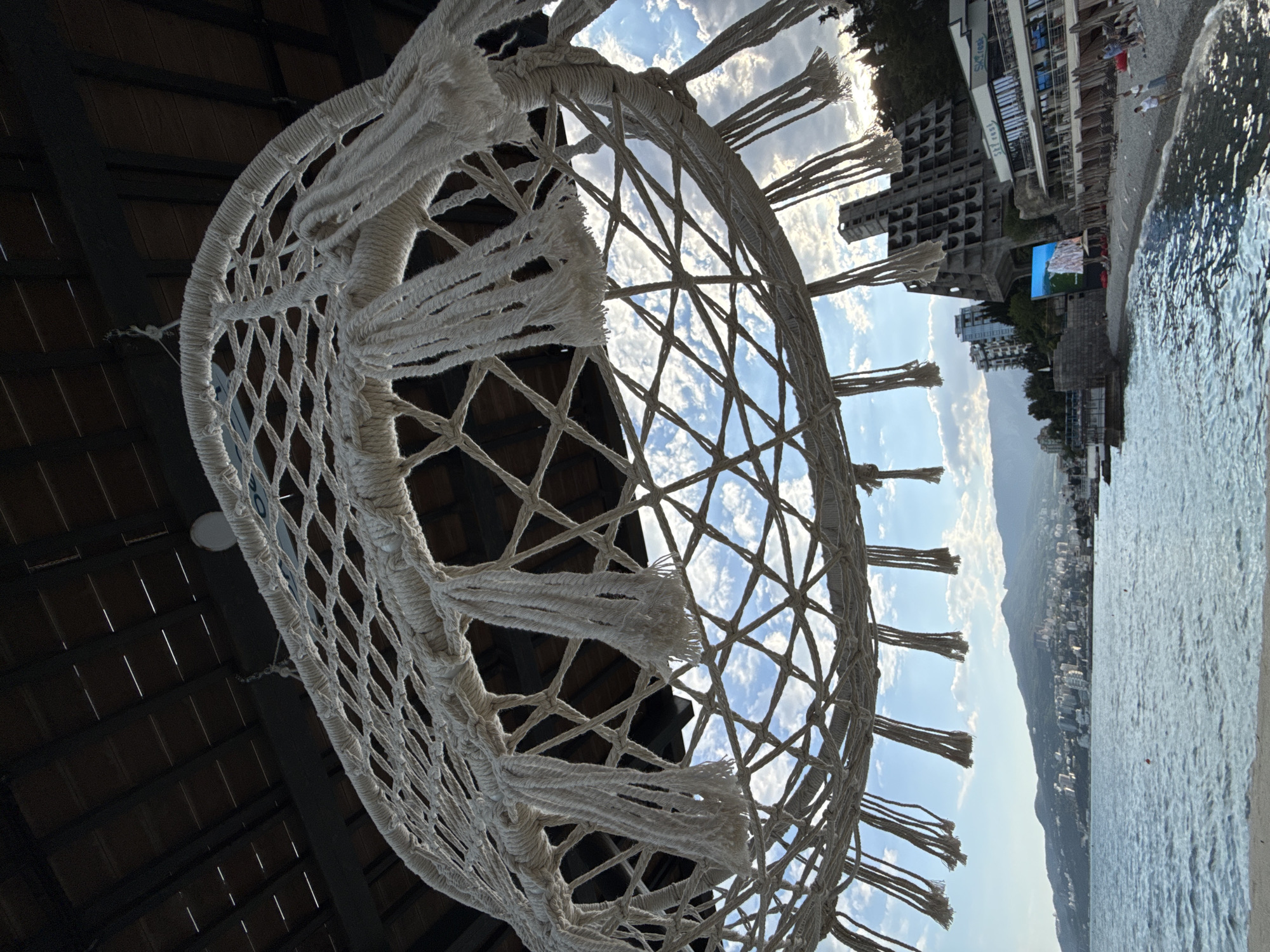 A low-angle, close-up shot focuses on a macrame hammock hanging from a dark, wooden roof. The hammock, made of thick, off-white rope, features a diamond-patterned net with tassels hanging from its lower edge. Through the hammock's open weave, a bright blue sky with scattered white clouds is visible, as well as a distant cityscape nestled against rolling green hills. The sea, a shimmering blue, laps at a pebble beach in the foreground. To the right of the frame, an unusual, brutalist-style concrete building with exposed structural elements looms over a modern-looking resort with a "Ski East" sign. Several beach chairs are set up on the shore, and a few people are walking or standing on the beach. The overall scene suggests a seaside resort in Yalta, Ukraine, on a bright, possibly late afternoon day.