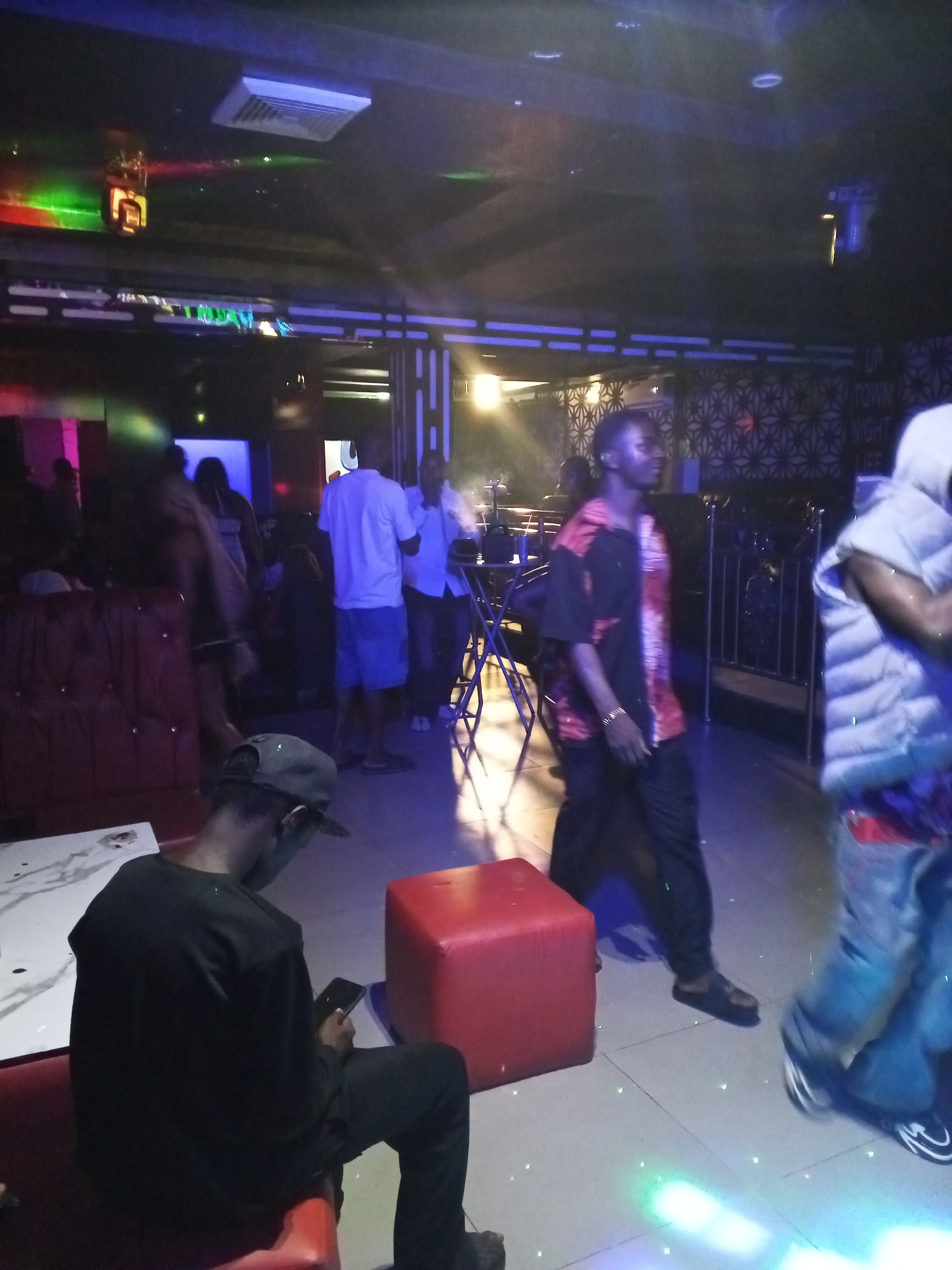 This image captures an indoor scene, likely a nightclub or lounge, at night in Adankolo, Nigeria. The lighting is dim and artificial, featuring vibrant blue, green, and red hues from various ceiling fixtures and spotlights that reflect on the tiled floor.
Several individuals are present, engaging in casual activities. In the foreground on the left, a person wearing a black long-sleeved shirt and a cap is seated on a red upholstered bench, looking down at a smartphone. A white table with a marble pattern is beside them. In the mid-ground, a person wearing a distinctive black and red patterned short-sleeved shirt and dark pants walks across the frame. To their right, another person, partially visible and blurred by motion, wears a white puffer vest over a colorful garment and blue jeans, also in motion.
In the background, more patrons are visible. A group of men stands near a small table and stools; one wears a white t-shirt and blue shorts, while another is in a white collared shirt. On the far left, red cushioned booth seating is visible, with blurry figures in the vicinity.
The venue features decorative elements, including a wall with a geometric pattern on the right side. Above this pattern, faint text on a sign reads "UP TOWN NIGHT LIFE," providing a clear indication of the establishment's identity. Overhead, an air vent and several light fixtures are visible, contributing to the club-like ambiance. The overall atmosphere appears relaxed and social.