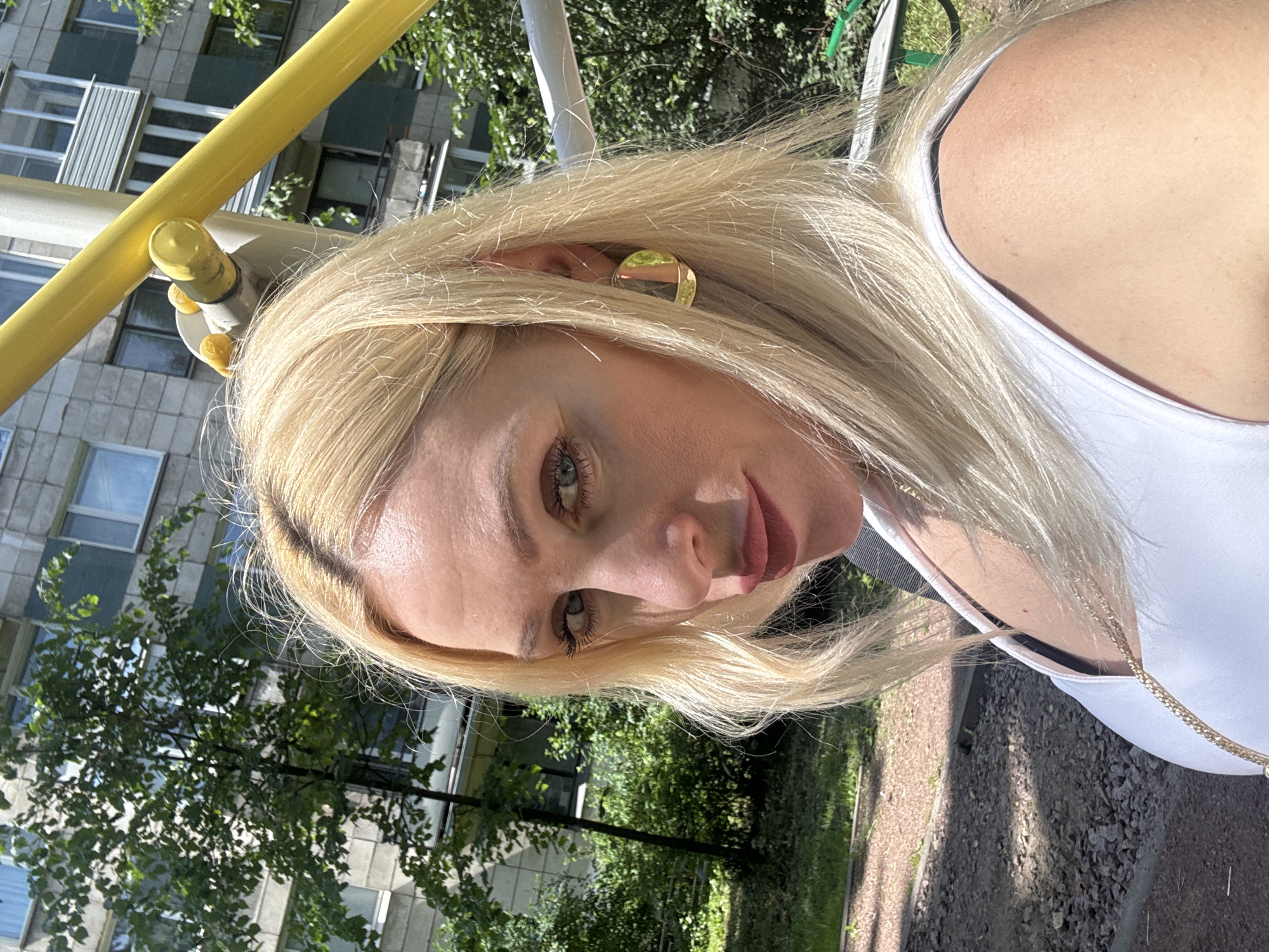 A woman with blonde hair and red lipstick poses for a selfie outdoors. She is wearing a white tank top and a gold hoop earring. Behind her, a multi-story building with balconies is visible, along with trees and a yellow structure that appears to be part of an outdoor gym or playground. The ground is covered with gravel and some patches of grass.
