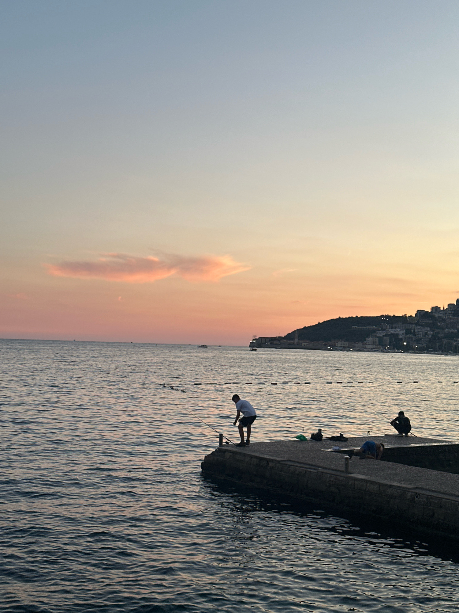An aerial view captures the sunset over the Adriatic Sea in Budva, Montenegro. The sky transitions from a soft, pale blue at the top to a warm orange and pink hue near the horizon, with a wispy pink cloud formation visible. The sea is calm, with gentle ripples reflecting the colors of the sky. A dark, man-made structure, likely a pier or breakwater, juts into the water from the right side of the frame. Several individuals are present on this structure. One person, wearing a light-colored t-shirt and dark shorts, stands and appears to be fishing. Another person, in a dark silhouette, is crouched down, also seemingly engaged in fishing. A third person is lying down on the structure. Several fishing rods are visible, and various objects, possibly fishing gear, are scattered on the pier. In the distance, a coastline is visible, featuring a hill covered with buildings, characteristic of Budva's urban landscape. The water is marked with a line of buoys. The overall scene depicts a peaceful evening by the sea.