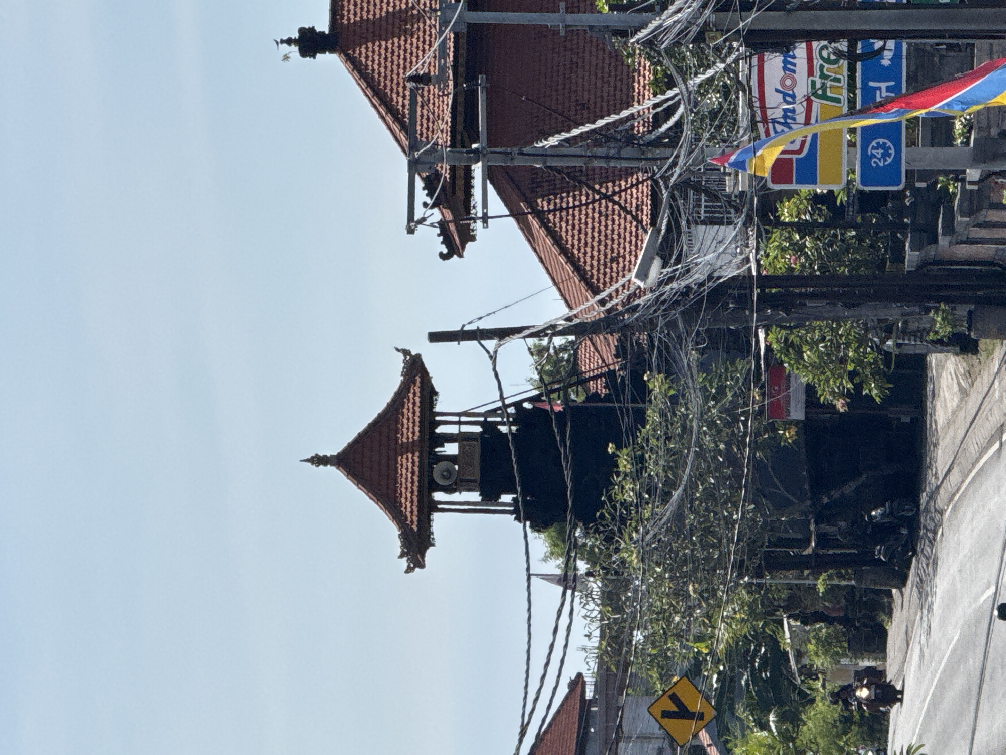 The image depicts a street scene in Kutuh, Indonesia, taken during the daytime. The sky is clear and light blue. A traditional Balinese structure with a tiered roof, possibly a temple or community hall, stands on the left side of the frame. Power lines are strung haphazardly between utility poles. On the right side of the image, commercial signs for "Indomaret" and "First" are visible, indicating the presence of a convenience store or a similar business. A triangular yellow street sign with a black arrow pointing left is present on the lower right. A road is visible on the right, with a motor bike driving away. Green foliage and trees fill the spaces in between buildings. The overall impression is of a typical busy street in a small town in Indonesia.