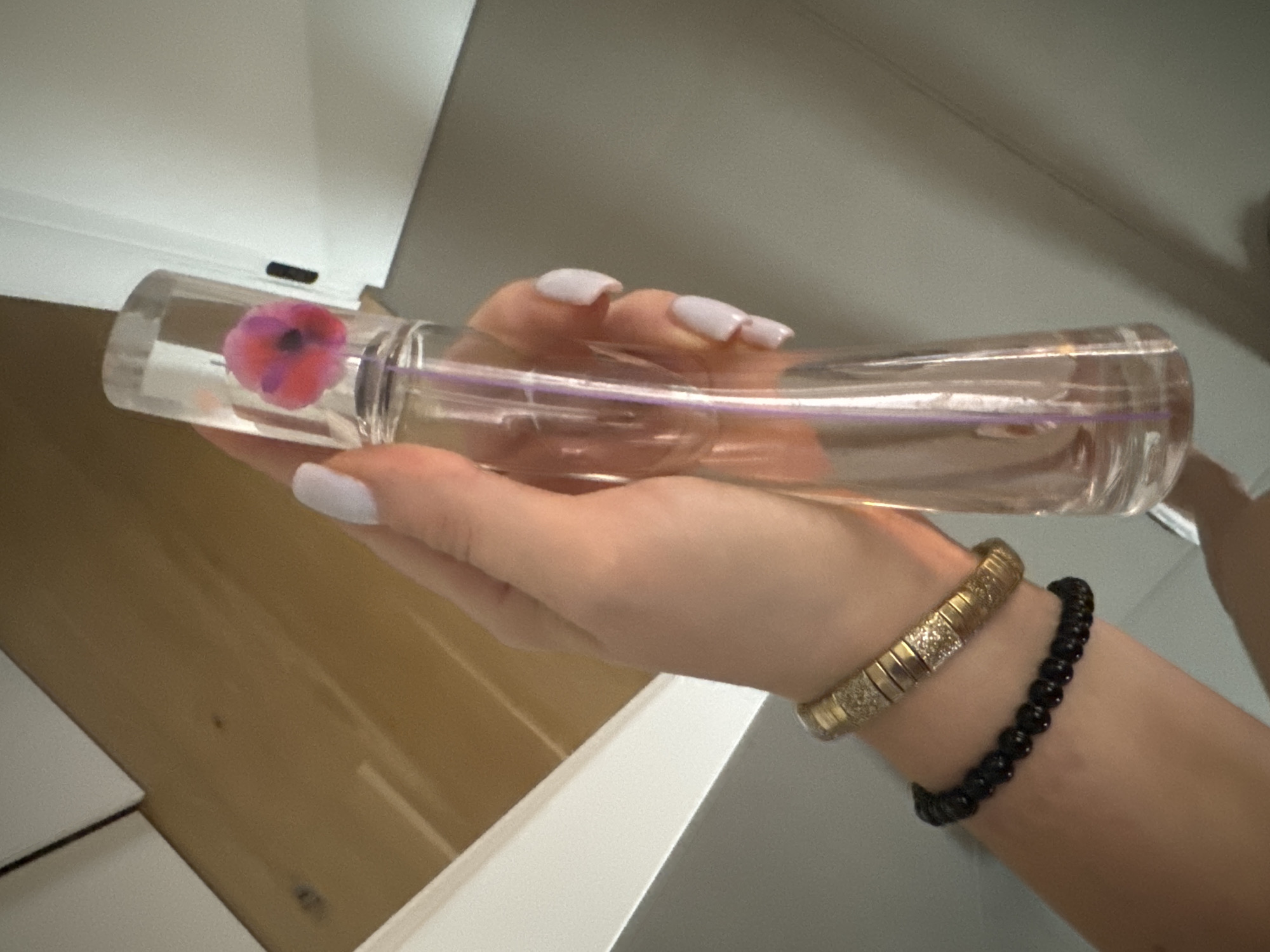 A person's hand, adorned with a light purple nail polish and two bracelets (one gold with small diamonds and one black beaded), holds a clear glass perfume bottle with a pink and purple flower design inside. The bottle has a purple liquid visible within. The background suggests an indoor setting with a wooden surface and a gray wall. There is no discernible location in the city of Murcia, Spain.