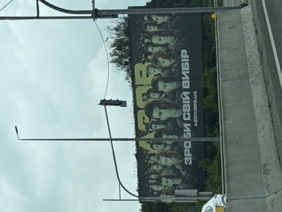 A billboard displays many portraits of people, presumably soldiers, against a black background. The word "AZOV" is prominently featured in yellow letters. Below, in Ukrainian, it reads "ЗРОБИ СВІЙ ВИБІР" (Make Your Choice) and "AZOV.ORG.UA". The billboard is positioned alongside a road, with traffic lights and street lamps nearby. The sky is overcast, suggesting a cloudy day.