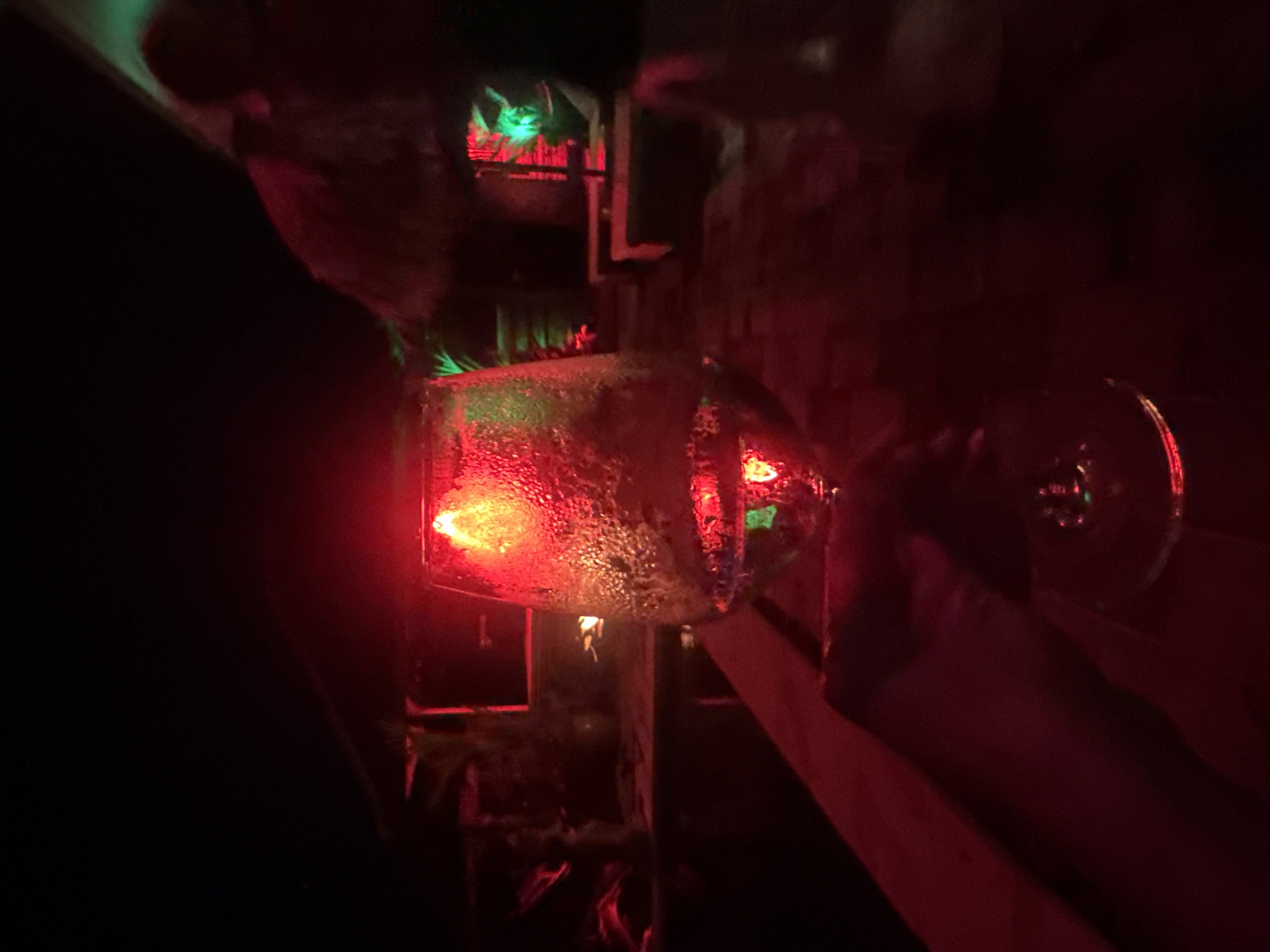 In the dimly lit location of Ungasan, Indonesia, a hand extends to hold a glass illuminated by a red light. The glass is covered in droplets, likely condensation. The background reveals a bar setting, with shelves visible, accentuated by both red and green lighting. The hand appears to be holding the glass towards the viewer. The overall atmosphere is dark and suggestive of a nightlife setting.