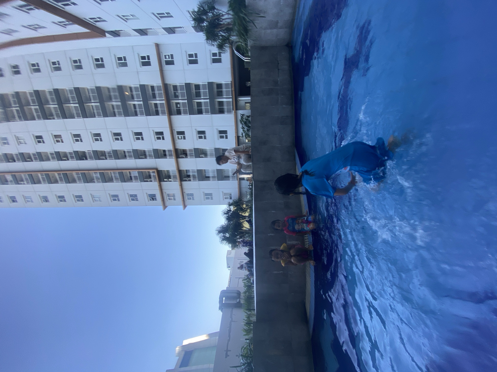 A low-angle, wide shot captures a swimming pool area with a large, modern apartment building in the background. The sky is a clear, bright blue. The apartment building is tall and white, with numerous balconies and windows, suggesting a residential complex. In the middle ground, a group of children are seen near the edge of the pool. Two young children are sitting on the pool's edge, one wearing a pink shirt with cartoon characters and the other a yellow shirt. Another child is partially visible, splashing in the water. A woman is seated on the edge of the pool, looking at her phone. Palm trees and other greenery are visible behind the pool area. The pool itself is filled with clear blue water, with ripples and splashes indicating activity. The overall scene suggests a leisure or recreational area within an urban or suburban environment, likely part of the housing development in Sidoarjo, Indonesia, given the architectural style and climate.