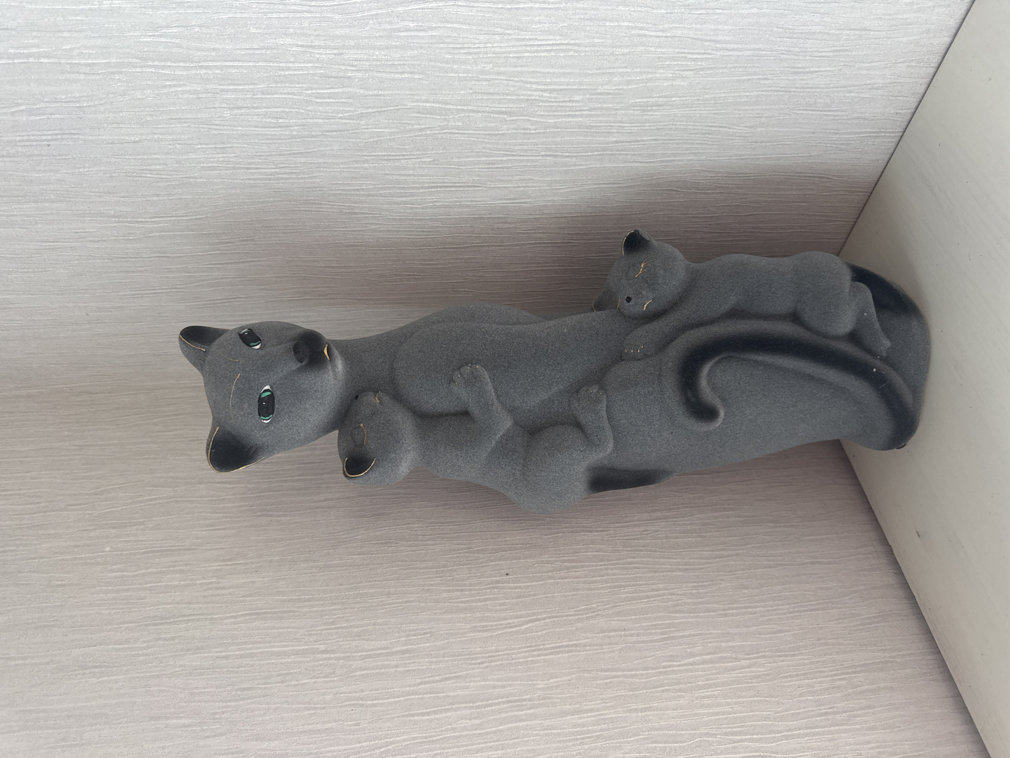 A gray, matte-finished ceramic sculpture of a mother cat with two kittens is placed on a white surface, likely a shelf or table, in the corner of a room. The mother cat is depicted standing, with one kitten clinging to her side and another nestled near her base. The sculpture has subtle gold accents on the ears and snout of the mother cat, and the eyes of the mother cat are painted green. The background consists of a light gray, textured wallpaper with vertical lines. The scene is lit from above, casting a soft shadow. The environment appears to be indoors. There are no people present in the image. The location is identified as Nikolaevka, Russia.