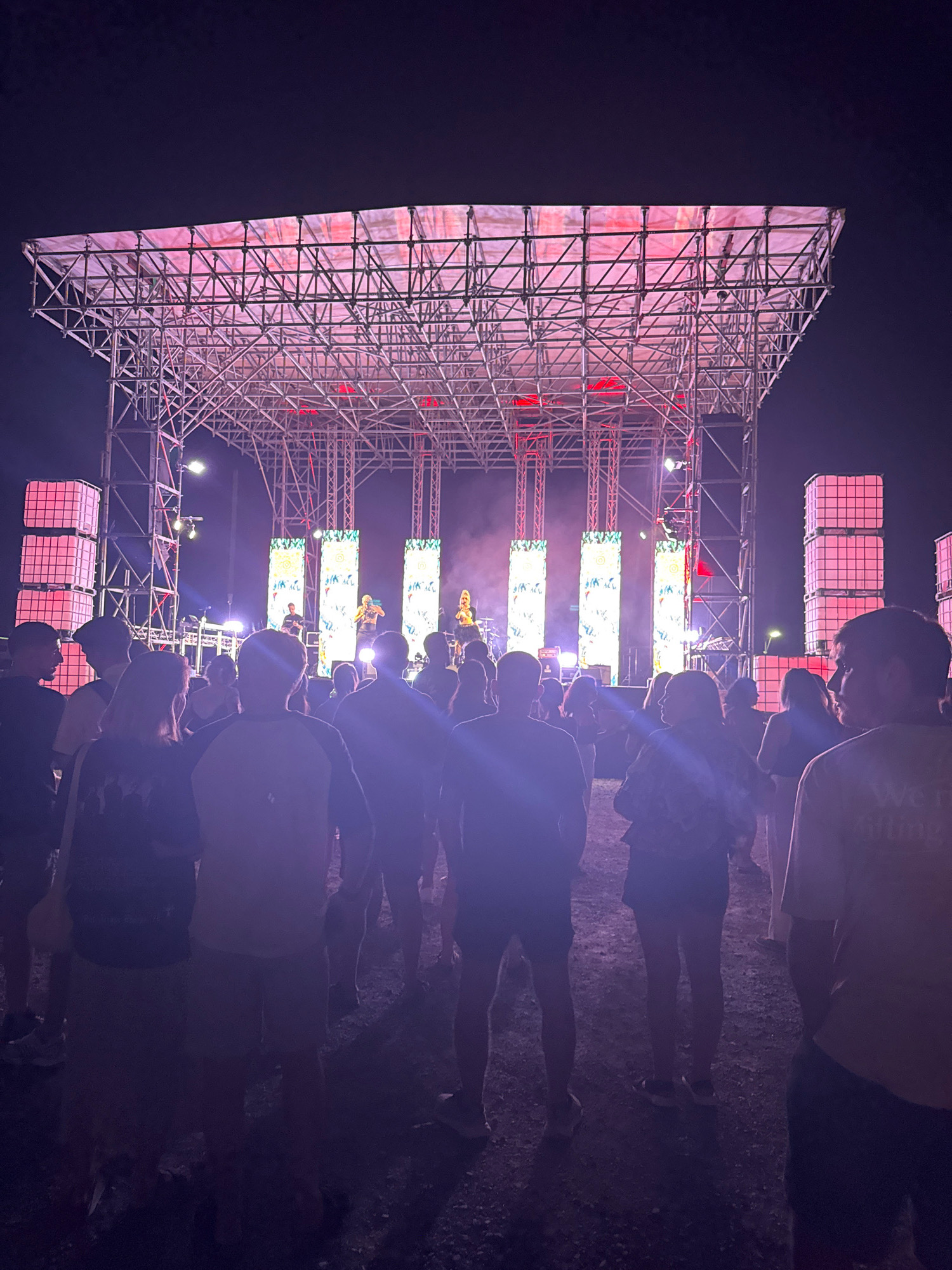 A crowd of people stands facing a brightly lit stage at an outdoor concert venue at night. The stage is constructed of metal scaffolding and covered with a translucent fabric illuminated from behind with pink and red lights. Three tall, narrow LED screens display colorful graphics. The crowd is silhouetted against the stage lights, with the foreground showing the backs of several individuals. To the left and right of the stage, large, translucent, pink-lit cubic structures are visible, possibly part of the stage setup or lighting infrastructure. The ground appears to be gravel or dirt. The overall atmosphere suggests a lively outdoor event.