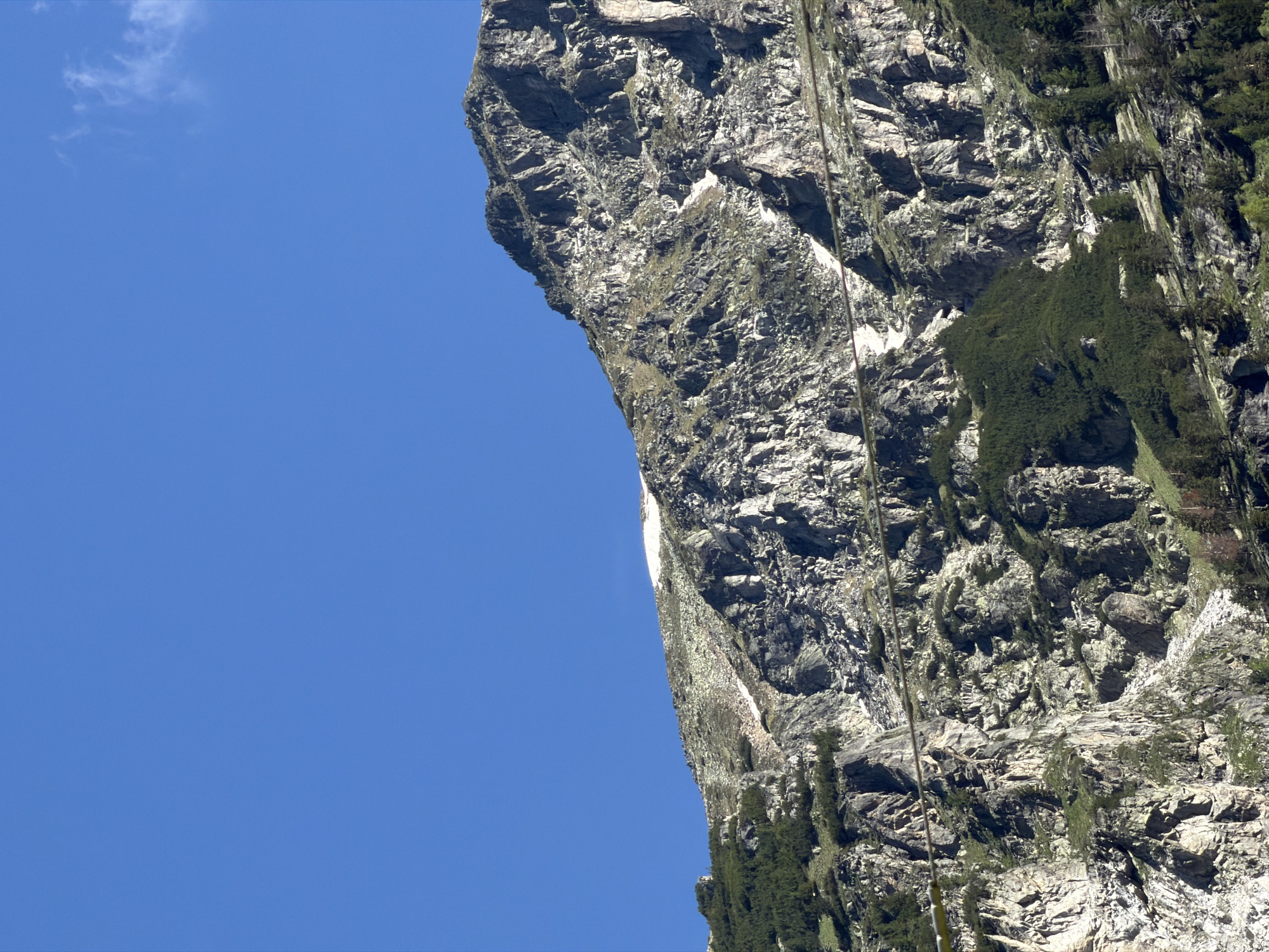 The image depicts a mountainous landscape with a clear blue sky. The lower portion of the image shows rugged, rocky slopes covered with patches of green vegetation, including trees. There are visible remnants of snow on some of the higher rocky areas. A thin, dark line, possibly a cable or wire, stretches horizontally across the middle of the frame. The overall scene is natural and unpopulated, suggesting a remote or wilderness area. The location is identified as Govedartsi, Bulgaria, a town known for its proximity to the Rila Mountains, which is consistent with the visual elements of the image.