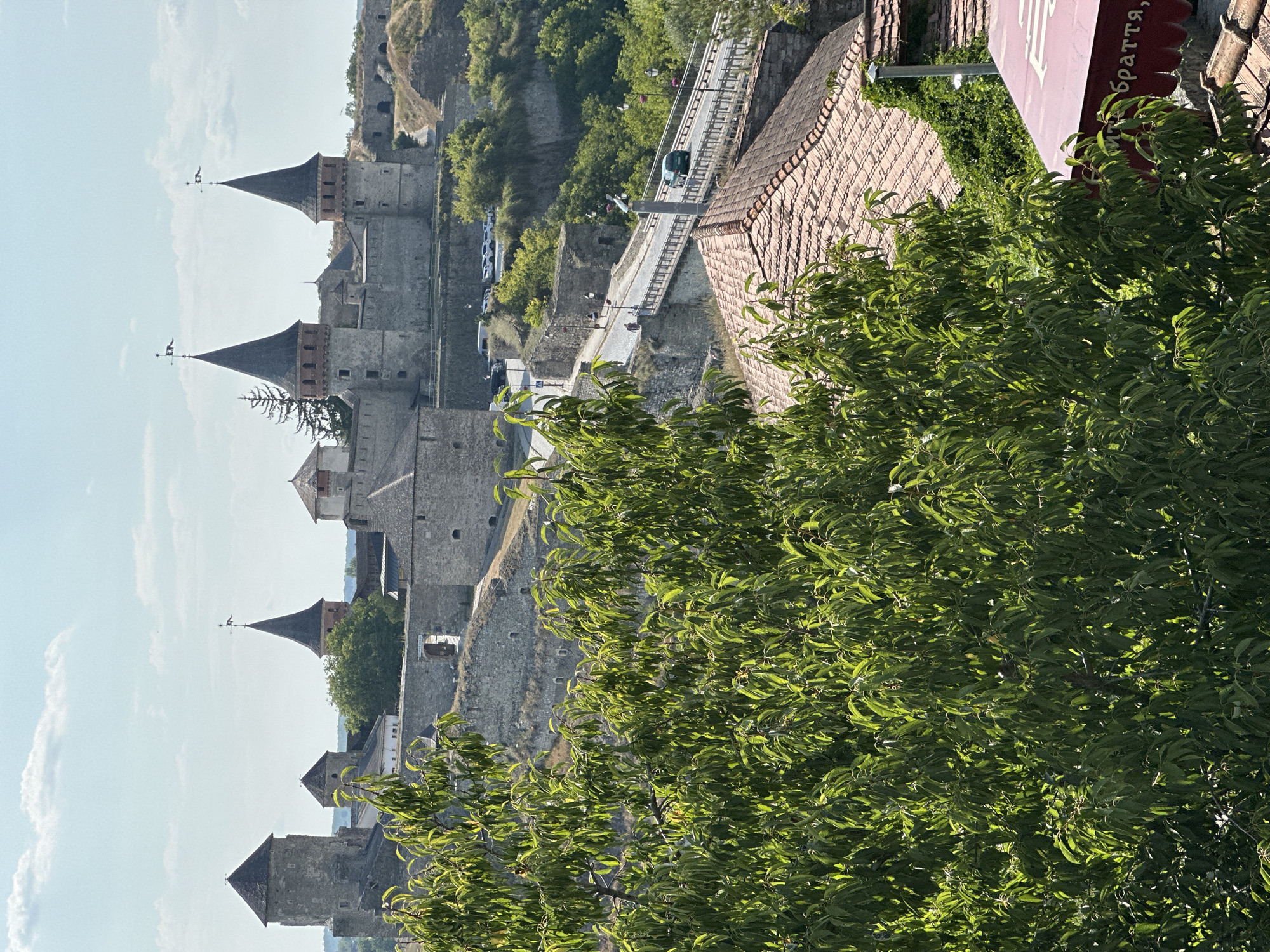 The image captures a section of the Kamianets-Podilskyi Castle in Kamianets-Podilskyi, Ukraine, on a clear day with scattered clouds. In the foreground, a lush green tree with dense foliage partially obscures the view. Below the tree, the terracotta-tiled roofs of several buildings are visible, one of which features a maroon awning with white Cyrillic text that reads "браття," meaning "brothers."
Beyond the foreground, the castle complex dominates the mid-ground and background. The ancient stone fortress features multiple towers with conical roofs, battlements, and weathered stone walls. A road winds its way towards the castle, crossing over a bridge. Several cars are parked near the castle entrance, and a few people are walking on the road and near the castle. The castle is situated on a rocky, elevated terrain, with green vegetation growing on the slopes surrounding it. The overall scene depicts a historic landmark bathed in sunlight.