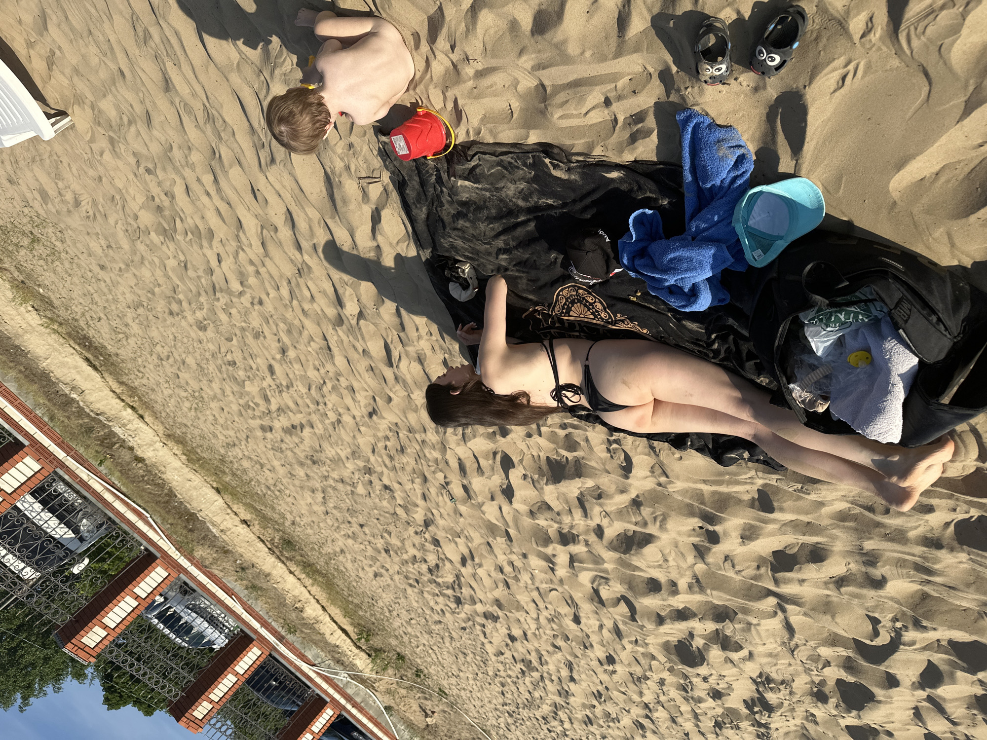 The scene is a sandy area, possibly a beach, located in Matiushino, Russia. A woman is lying on a black blanket wearing a black bikini. Beside her is a bag, a blue towel, and a light blue hat. Nearby, a child is playing in the sand with a red bucket. A pair of children's shoes are also present. The area appears to be near a building with a red brick and white facade and a wrought iron fence.
