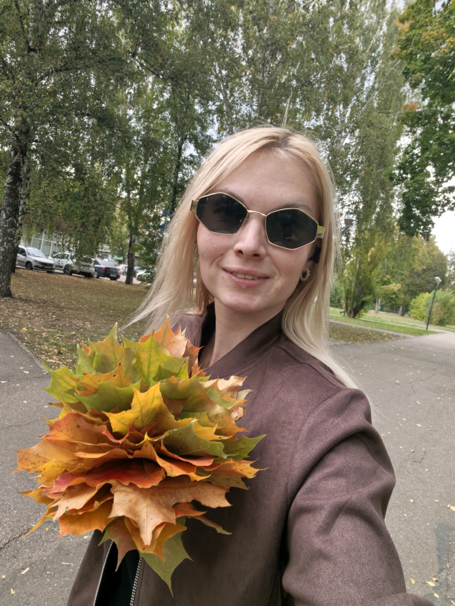 A blonde woman wearing sunglasses and a brown jacket holds a bouquet of colorful autumn leaves in a park-like setting. The leaves are a mix of green, yellow, orange, and red. In the background, there are trees with foliage, some cars parked on the side of a road, and a paved pathway. The scene appears to be in a city park in Sterlitamak, Russia, during autumn.