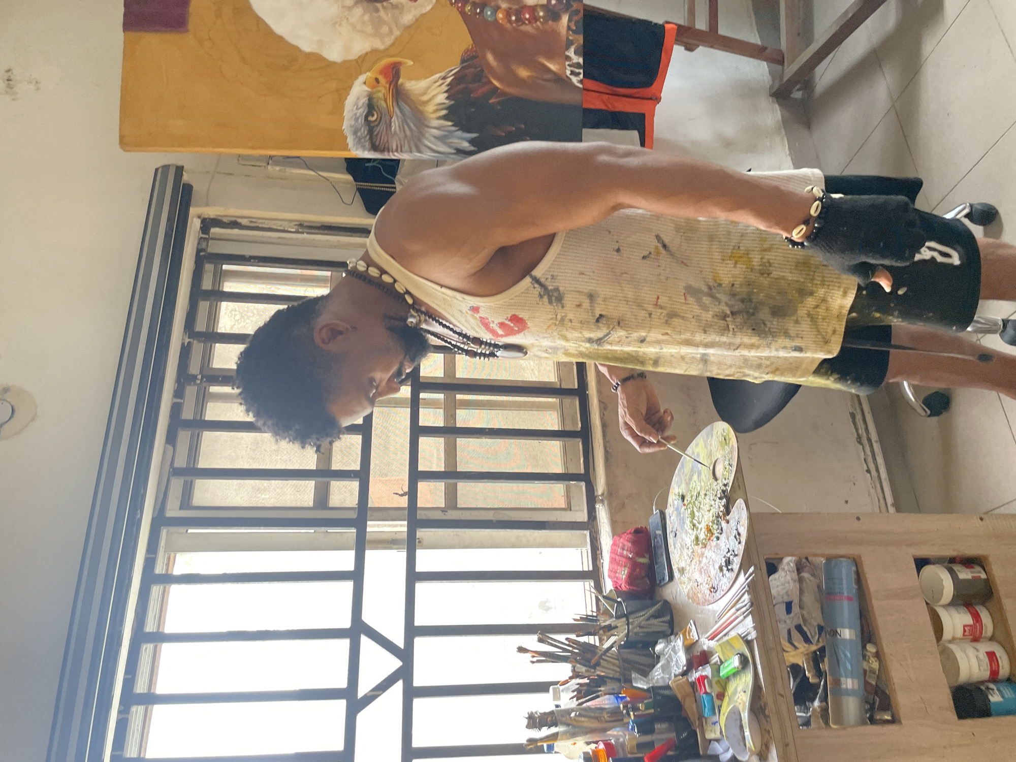An artist, a Black male with dark, curly hair and a beard, stands in his studio in Ado, Nigeria, engaged in painting. He wears a paint-splattered white tank top, black shorts, and a black fingerless glove on his left hand adorned with a bracelet made of shells and dark beads. Around his neck, he wears multiple necklaces, including one with a large, dark pendant and another with a string of brown beads. He is holding a paintbrush, mixing paint on a wooden palette on a desk.
The desk is cluttered with art supplies, including numerous paintbrushes in holders, bottles of paint, tubes of paint, and other art materials. Behind the desk is a window with metal bars. To the right of the artist is a large canvas painting featuring the head and shoulders of a majestic eagle, its piercing gaze directed towards the viewer. Partially visible next to the eagle is another painting or canvas, showing a section of a person's shoulder and neck adorned with a colorful beaded necklace, suggesting a portrait in progress. The studio appears to be a personal workspace, with art supplies and works in various stages of completion.