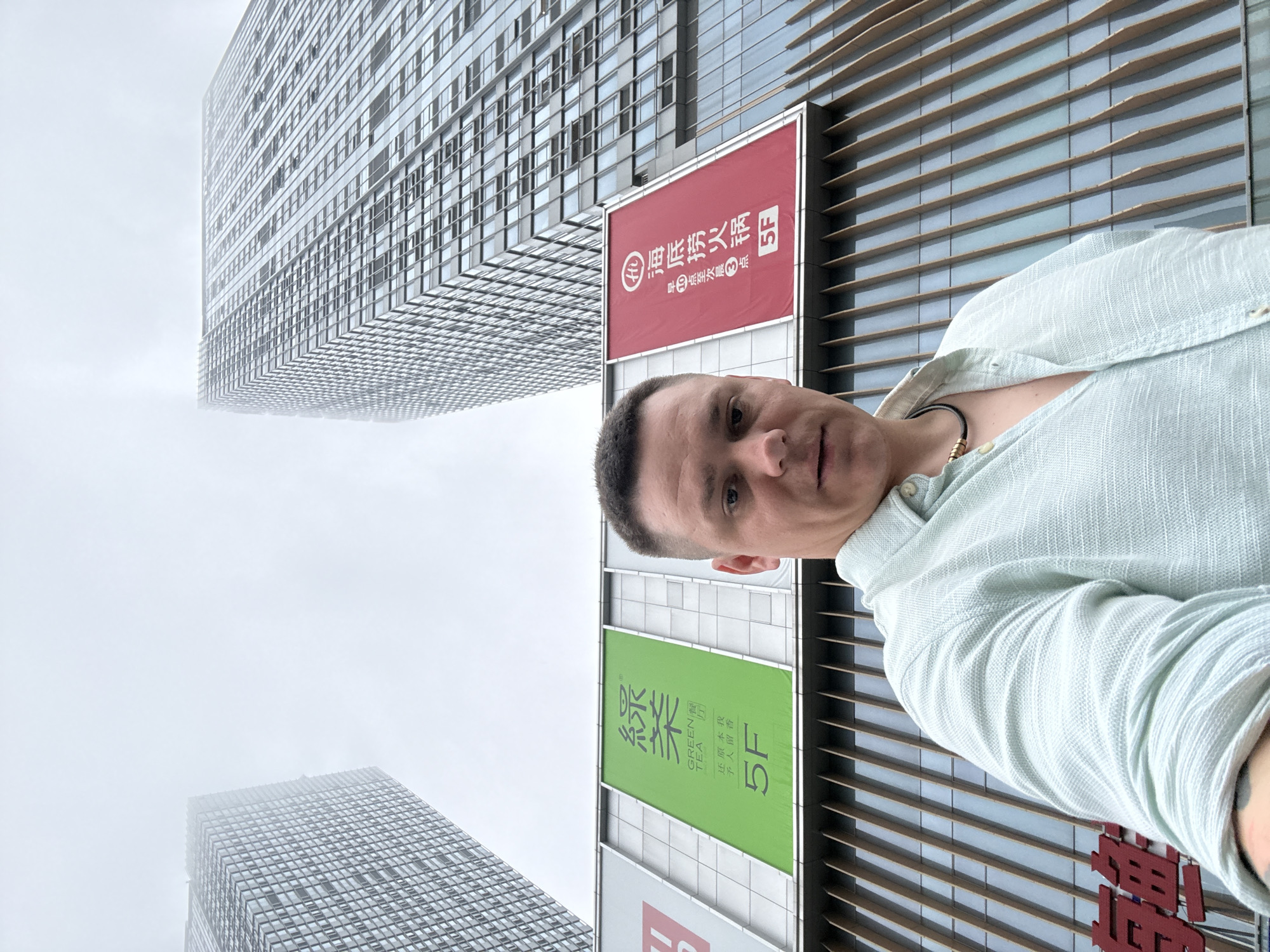 A low-angle, full-frame, eye-level selfie is taken from a man's perspective in an urban environment. The man, appearing to be of Caucasian descent, has short brown hair and is looking directly at the camera. He is wearing a light blue, short-sleeved, textured button-down shirt, open at the collar, revealing a dark necklace with gold accents.
Behind the man, two tall, modern glass-clad skyscrapers rise into a hazy, overcast sky. The buildings are characterized by their grid-like window patterns. In the mid-ground, a horizontal banner hangs between the two skyscrapers. This banner is divided into two sections. The left section is bright green with Chinese characters and the text "GREEN TEA" along with "5F". The right section is red with Chinese characters and the text "5F".
At the bottom of the image, part of another building facade is visible, featuring vertical wooden or wooden-effect slats over glass. A large red sign with Chinese characters is partially visible at the bottom left of the frame. The overall scene suggests an outdoor setting in a city, possibly a commercial district, with the hazy sky indicating a cloudy or foggy day.
Based on the prominent signage for "GREEN TEA" (綠茶) and "Hai Di Lao Hot Pot" (海底撈火鍋) with the "5F" designation, and the architectural style, the location is identified as the Wanda Plaza in Qinhuangdao, China. Wanda Plaza is a common commercial complex in many Chinese cities, and the specific signage points to this particular establishment.