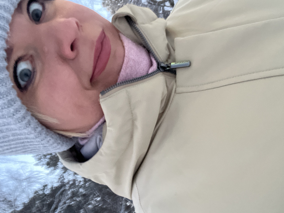 A woman's face, slightly distorted, with large blue eyes and pink lips, is positioned in the upper left corner. She wears a gray knit hat and a pink sweater. The rest of the image is dominated by a tan, quilted jacket. The ground, with snow and possibly branches, is visible in the lower-left corner. The zipper of the jacket is partially visible.