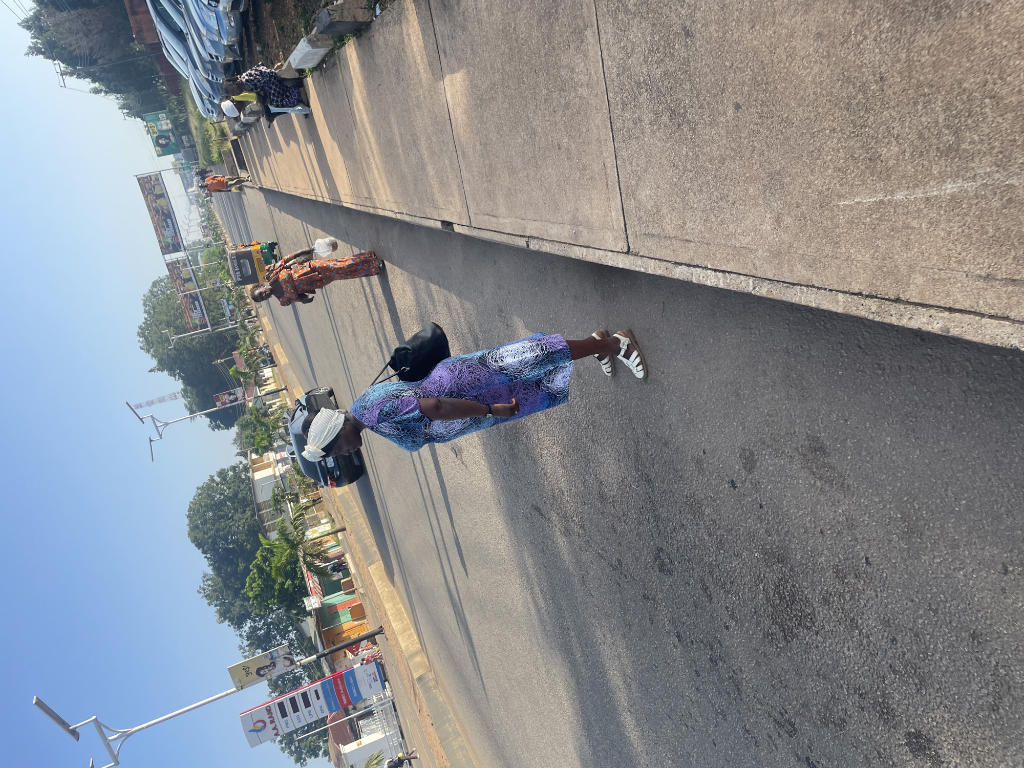 This image depicts a street scene in Jos, Nigeria, likely during daytime. The foreground is dominated by a woman wearing a vibrant purple and blue patterned dress and a white headscarf, standing on the asphalt road. She is facing to the right of the frame, with a black handbag slung over her shoulder. Her white sandals are visible. Behind her, on the sidewalk, another woman in an orange and patterned dress walks away from the camera, carrying a white bucket. Several other individuals are visible further down the street, including a man in an orange shirt walking on the sidewalk and two individuals sitting on the curb. Cars are parked along the right side of the street, and a three-wheeled vehicle, possibly a Keke Napep, is visible in the distance on the road. A gas station with prominent signage is located on the left side of the street, alongside various shops and buildings. The sky is clear and blue. The overall impression is of a bustling urban environment with everyday activity.