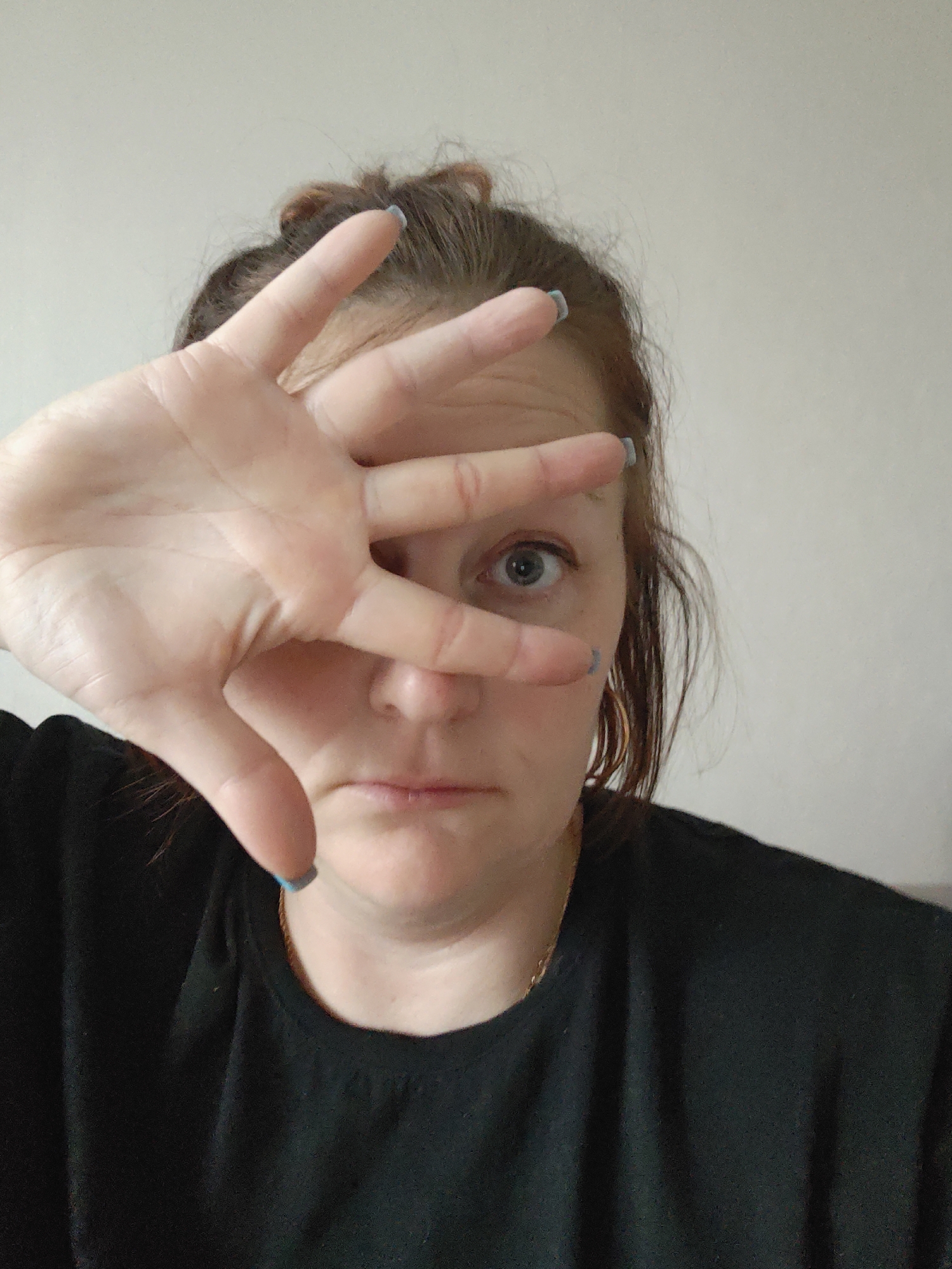 A woman with reddish-brown hair pulled up in a messy bun is facing the camera, with her right hand held up in front of her face. She is wearing a black t-shirt and gold hoop earrings. Her fingernails appear to be painted a light blue color. Her eye is visible between her fingers. The background is a plain, light-colored wall. There is no information about Markovskii, Russia.