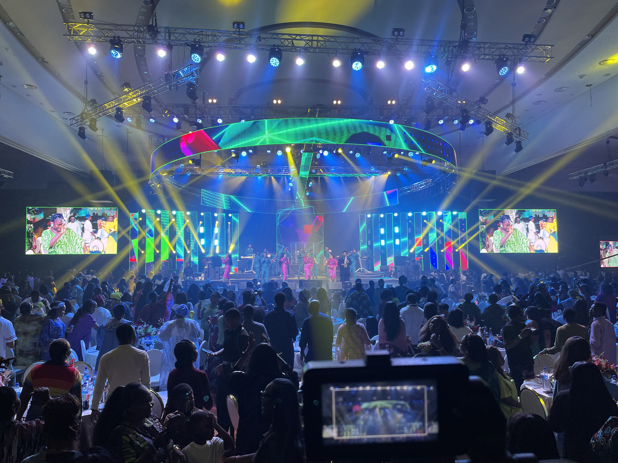 Captured from a ground-level perspective, the image presents a lively indoor event, likely a concert or celebration, in an unidentified city. The scene is dominated by a large crowd of attendees, most of whom are facing towards a stage. The stage is elaborately lit with spotlights and features a circular truss structure. Performers are visible on the stage, dressed in various colors. Large screens flank the stage, displaying content that mirrors the on-stage activity. The room is well-lit, and tables with floral centerpieces can be seen among the crowd. In the foreground, a camera rig is noticeable, suggesting the event is being recorded.