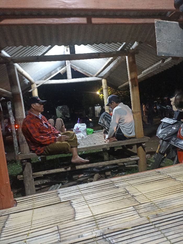 The image depicts an evening scene under an open-air structure in Kecamatan Pujut, Indonesia. The time is clearly night, illuminated by artificial lights casting shadows.
In the foreground, two men are prominently seated on a raised wooden bench, both barefoot. The man on the left wears a red and black plaid shirt, brown pants, and a dark baseball cap, looking towards the left side of the frame. The man on the right wears a light-colored long-sleeve shirt, striped lower garment (possibly a sarong or pants), and a dark baseball cap, looking down. Another person is partially visible behind the man in plaid.
The structure itself is constructed with bamboo poles and features a corrugated metal roof. Below the seating area, the ground appears to be dirt with some scattered debris. A bamboo-slatted platform or floor extends into the foreground, showing signs of wear.
Between the two main figures, on the bamboo platform, a white plastic bag displaying the text "TARA" and a green plastic bag are visible. To the far right, a dark-colored scooter motorcycle is parked, with part of its license plate visible, reading "DP 3710 Q" and "08 24". In the background, more people are dimly visible, suggesting a small gathering or communal space. The overall atmosphere appears relaxed and informal.