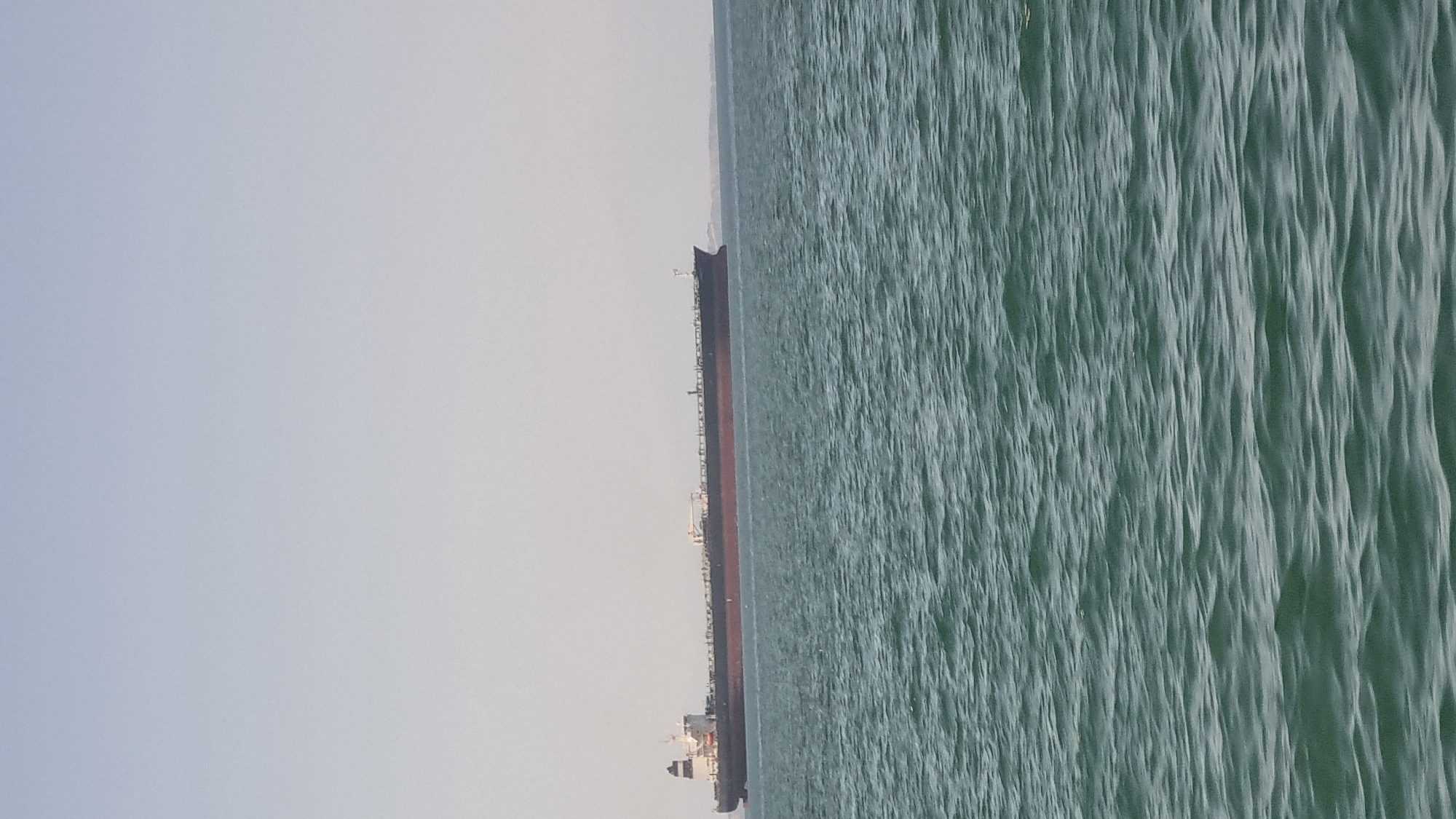 An oil tanker ship is shown sailing on the sea with a light blue sky in the background. The tanker is long and dark, with a red hull. The water is choppy and a deep green color. The sky is clear and hazy. There is a distant landmass on the horizon. The image is a still photograph taken from a boat or from shore. There are no people visible in the image. The location is the sea.
