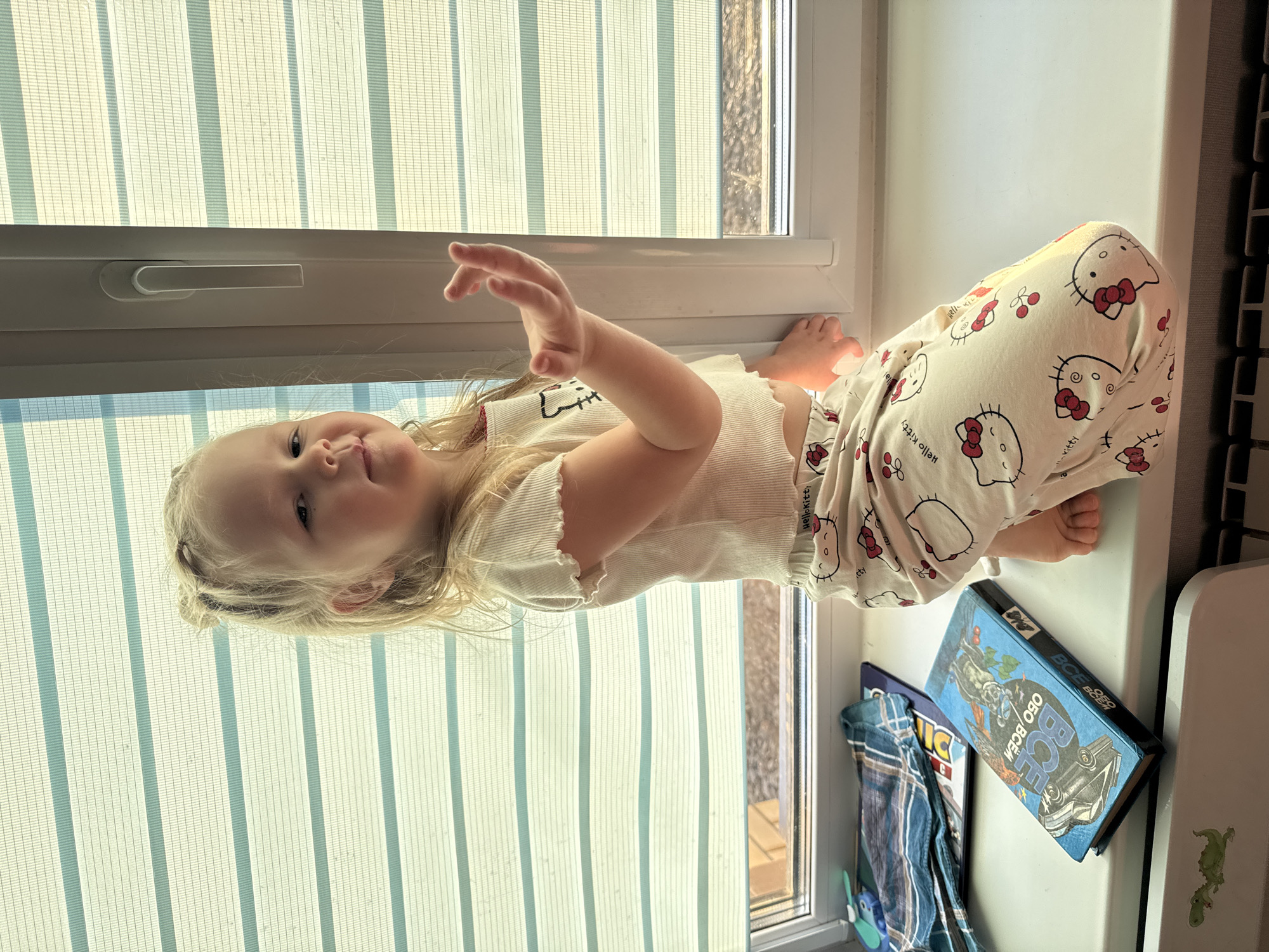 A young child with blonde hair is lying on a windowsill, wearing a white Hello Kitty pajama set. The child's body is positioned diagonally across the frame, with their head near the window's edge. The window is covered with vertical blinds. Next to the child is a stack of books. The image appears to have been taken indoors in Gornyi (Dvurechenskoe S/P), Russia.