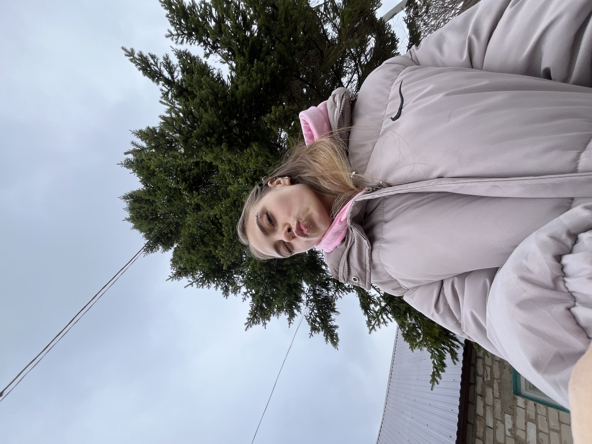 A young woman in a light-colored puffer jacket stands outdoors with a coniferous tree behind her. The location appears to be in Фоки, Russia, as indicated by the request. The sky is overcast. A building with a corrugated metal roof and brick walls is partially visible. The woman is looking at the camera with her lips pursed.