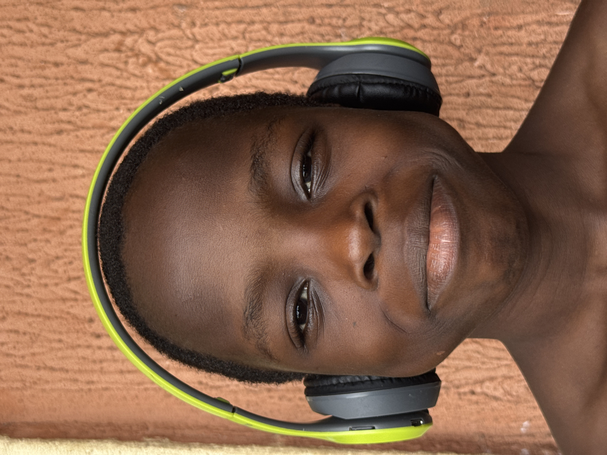 This is a close-up, rotated portrait of a young person with dark skin, positioned against a textured, reddish-brown background. The subject is wearing over-ear headphones, which are primarily dark grey with prominent lime green accents on the headband and ear cup rims. They are looking directly at the viewer with a calm and gentle expression, featuring a subtle, soft smile. Their dark eyes are open, and their short, dark hair is visible beneath the headphone band. The background exhibits an irregular, mottled pattern, suggesting a painted wall or concrete surface, consistent with building materials found in locations like Onitsha, Nigeria. The lighting is soft and even, highlighting the subject's features without harsh shadows. No specific time of day or weather can be inferred, and no visible text is present in the image.