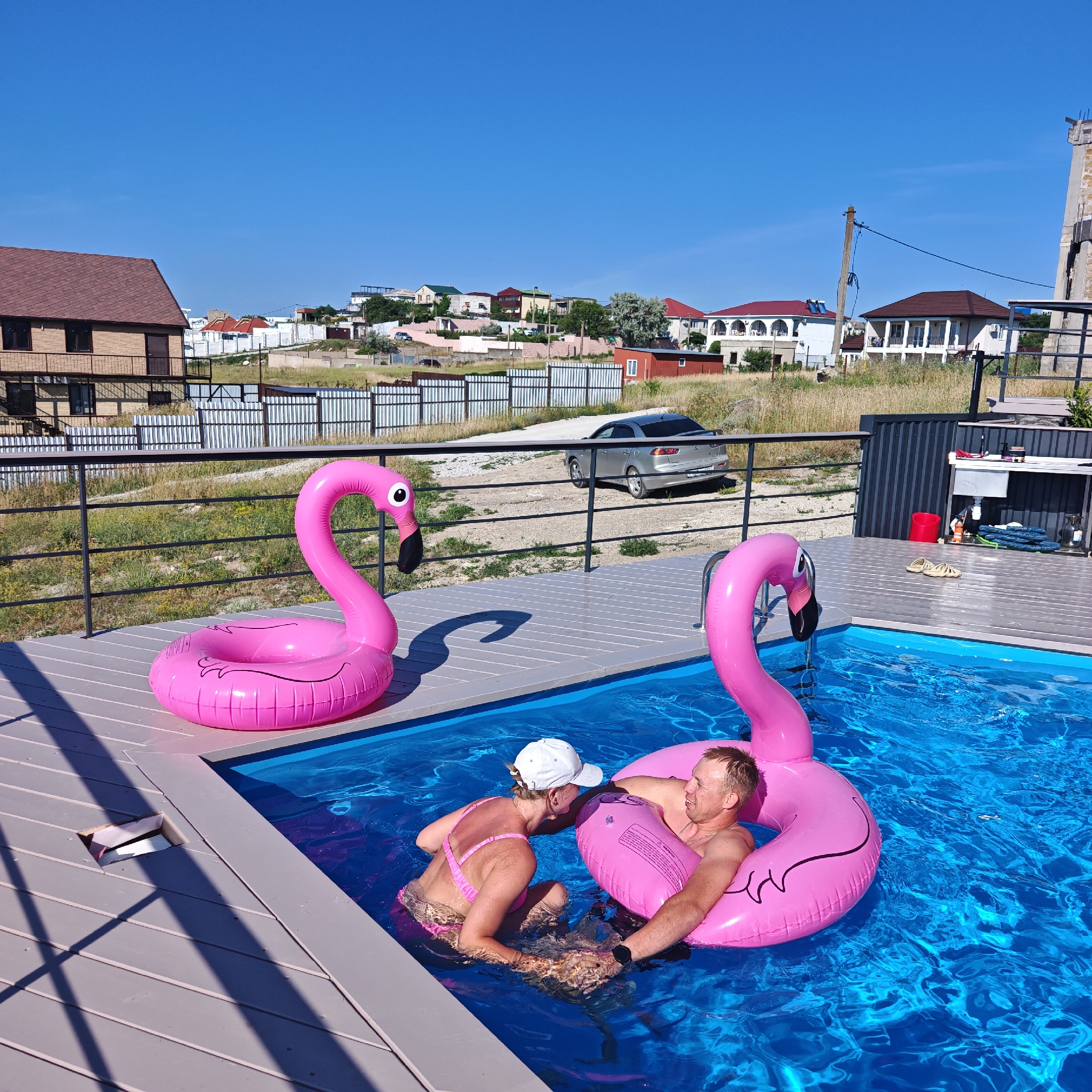 Two people are relaxing in a bright blue swimming pool on a sunny day. They are both using large, inflatable pink flamingo pool floats. The woman is wearing a pink bikini and a white baseball cap, and the man is wearing a watch on his left wrist. They are leaning against each other, appearing to be in a comfortable and friendly interaction.
The pool is surrounded by a grey wooden deck, and a black metal railing runs along the edge. In the background, there is a dry, grassy area with several houses, fences, and a silver car. The sky is a clear, deep blue, suggesting it is a warm and pleasant day. The overall setting appears to be a residential area, possibly a backyard or vacation property. There is some visible text on the inflatable flamingo floats, but it is too blurry to read. The scene evokes a sense of leisure and enjoyment.